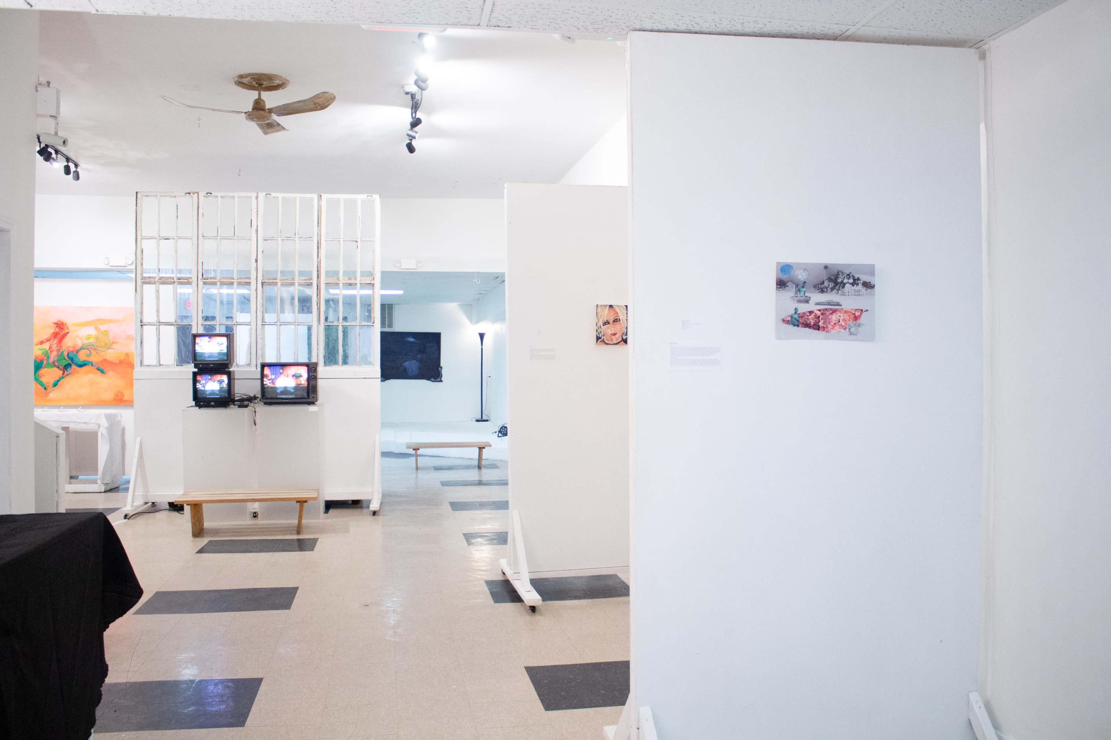 The image displays an art gallery with white walls, various artworks hung, two televisions on a stand, and a colorful painting in the background.