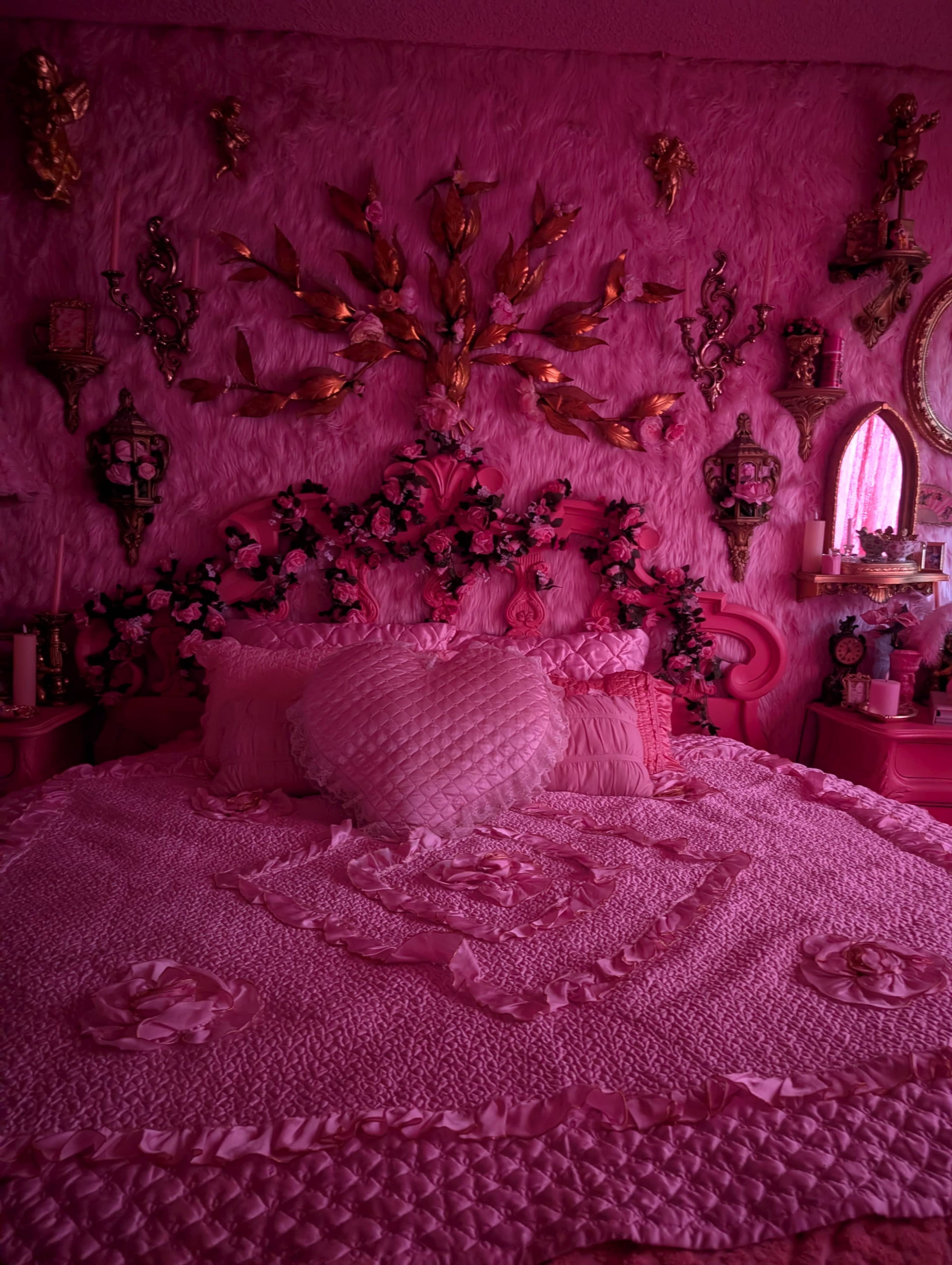 The image shows a bedroom with pink walls, a pink bedspread adorned with a heart-shaped pillow, and various decorative elements including wall art and floral arrangements.