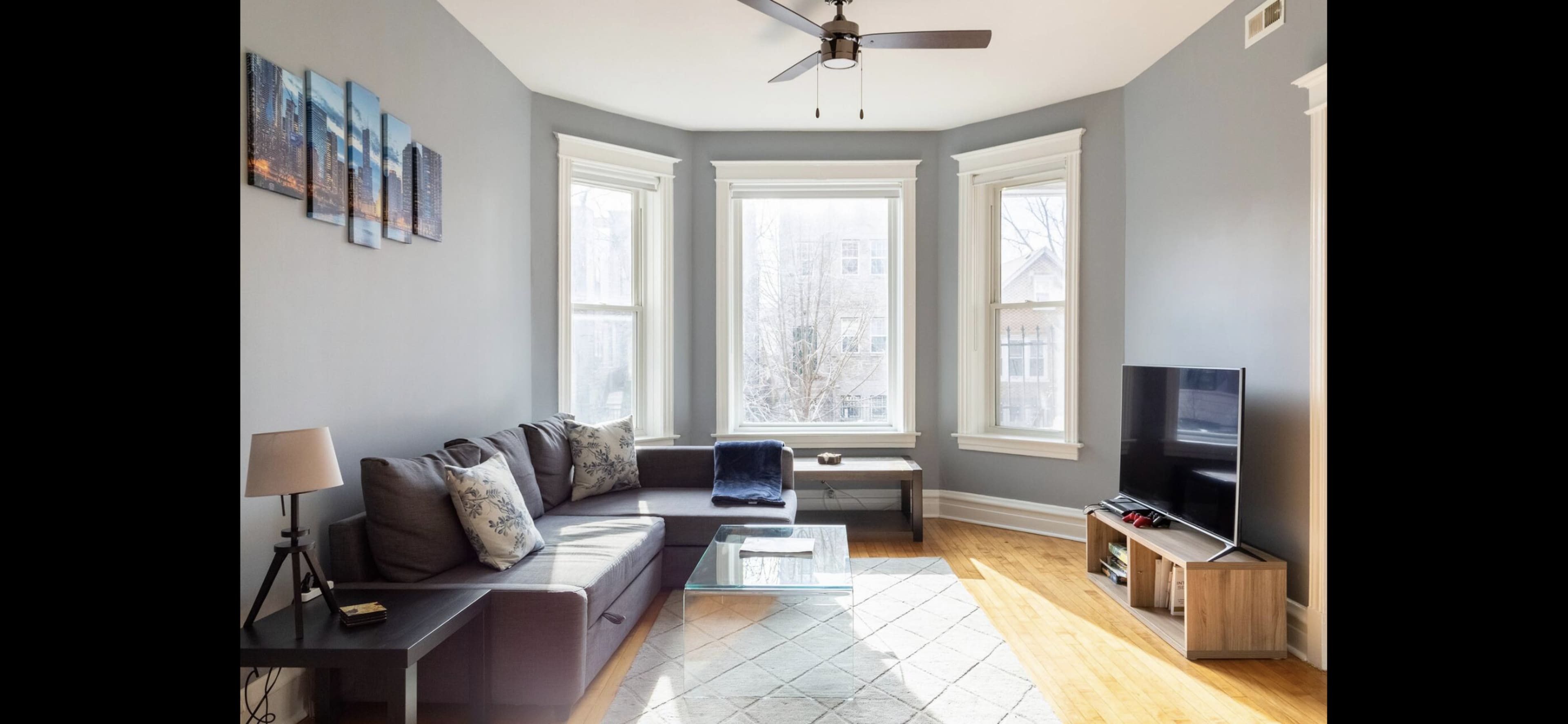 A modern living room with a sectional couch, a coffee table, and a television facing large bay windows that allow natural light to enter the space.