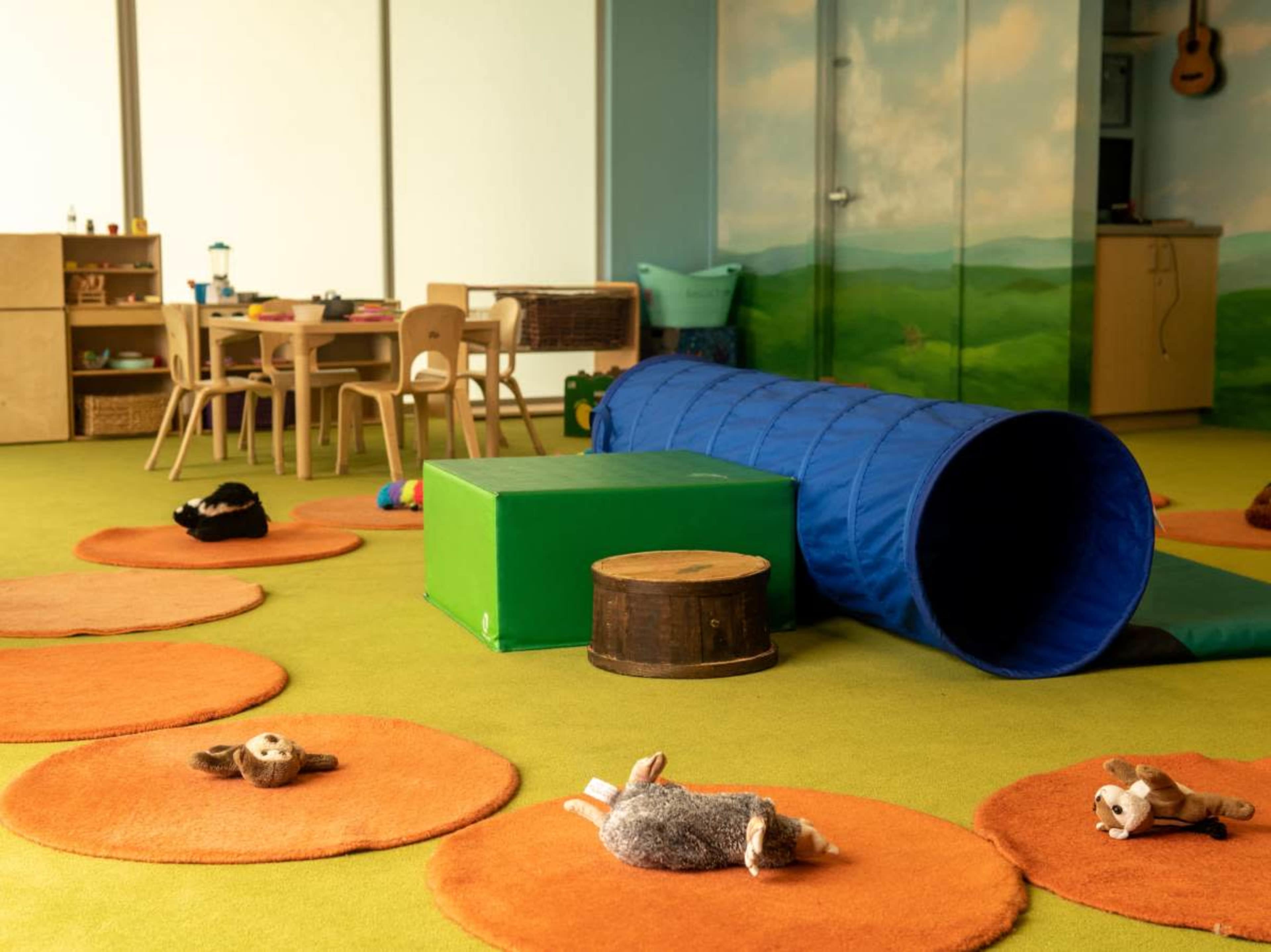 A colorful playroom with orange circular rugs, a blue tunnel, a green block, and plush toys scattered on the floor.