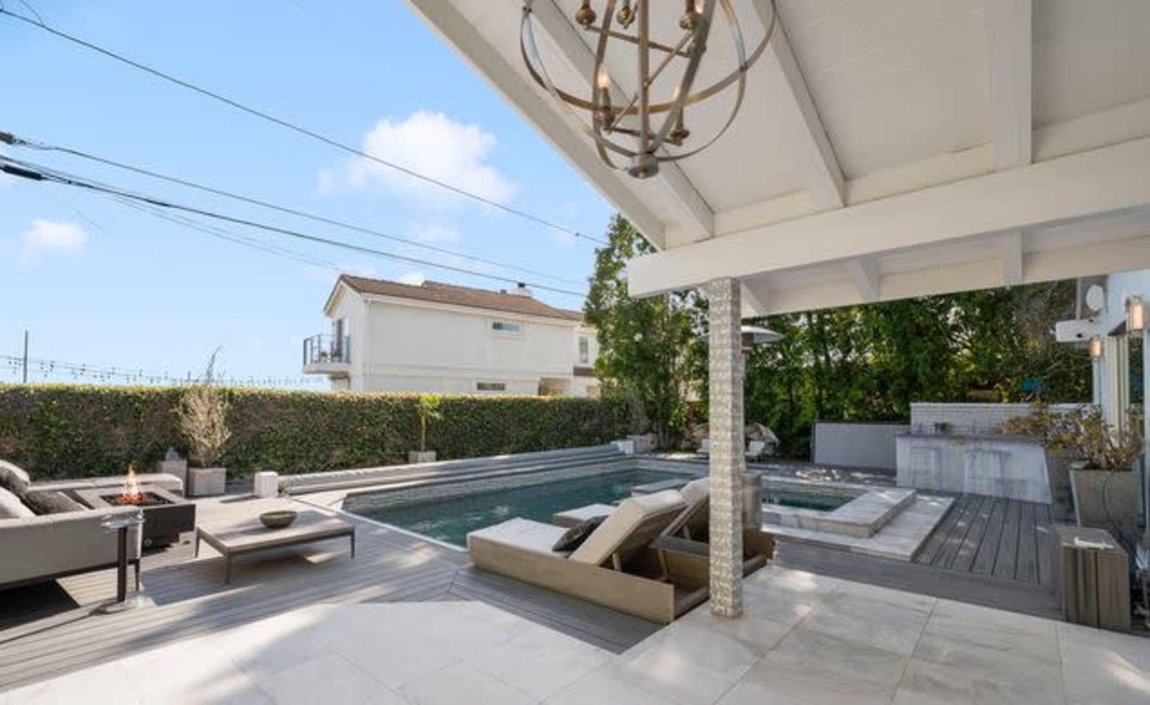 The image shows a sunlit outdoor patio featuring a swimming pool, a hot tub, and modern seating arrangements beneath a partly covered roof.