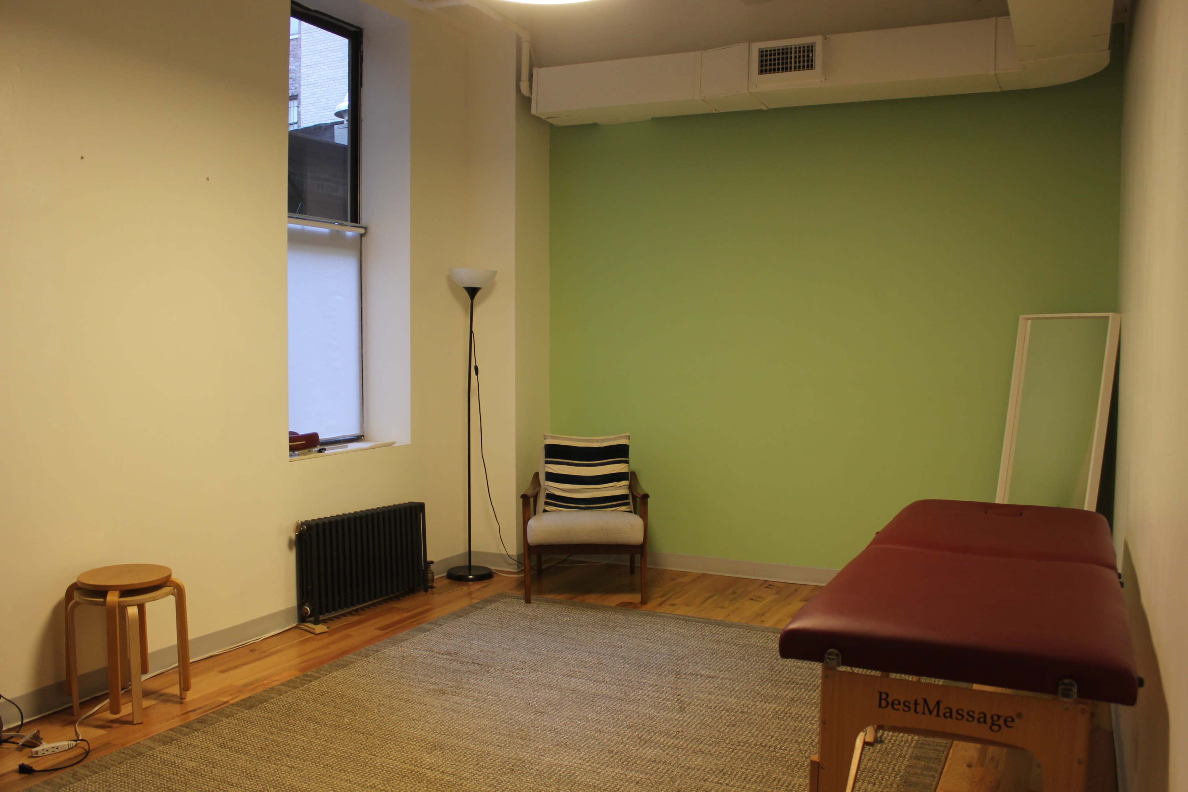 A therapy room with a red massage table, a chair, a stool, a lamp, and a window, all set against a green wall.