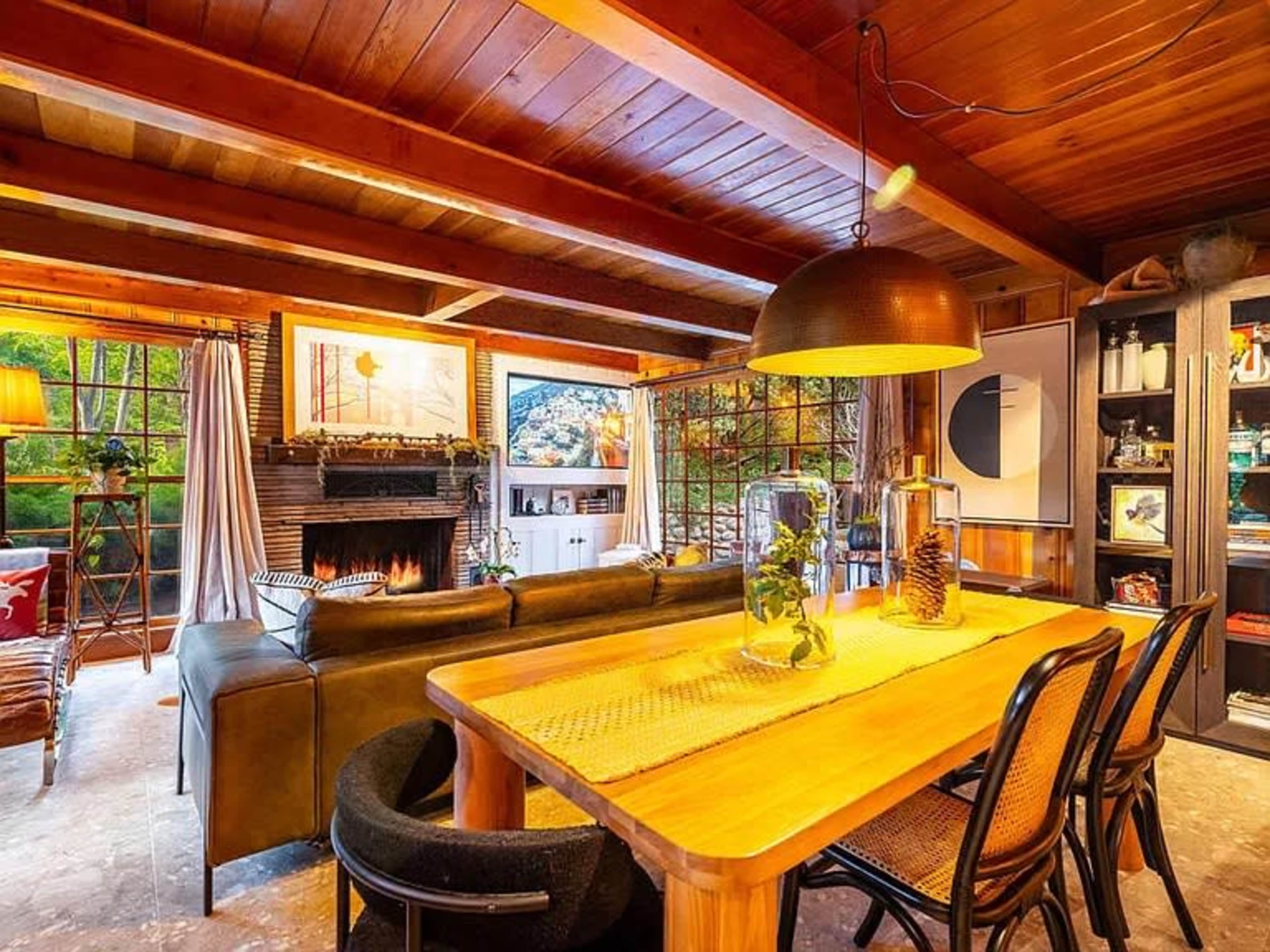 The image shows a cozy living area with a wooden ceiling, a fireplace, a dining table, and large windows allowing outdoor light to enter.