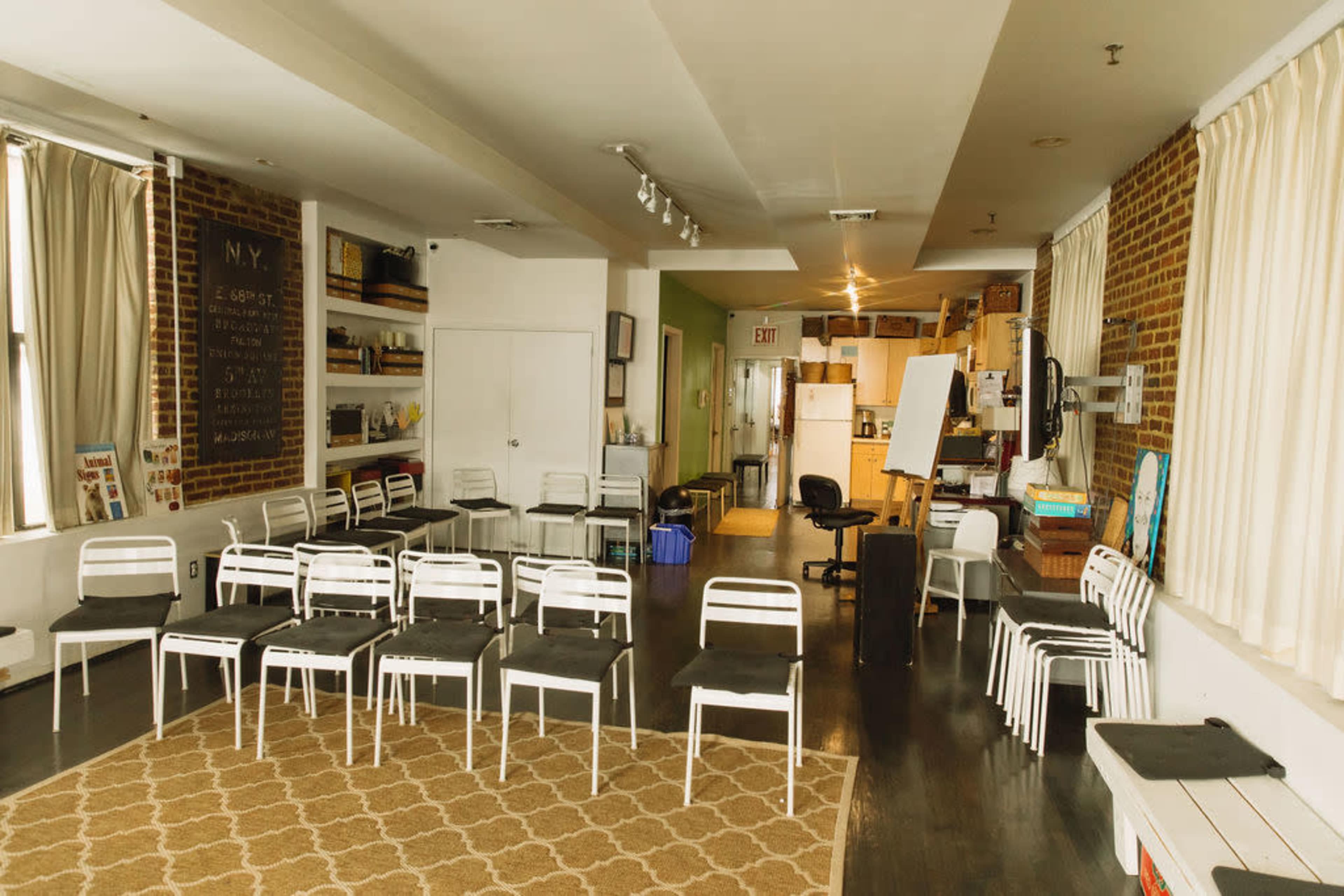 The image shows a spacious room set up with white chairs arranged in rows, a patterned rug on the floor, and various furnishings along the walls.