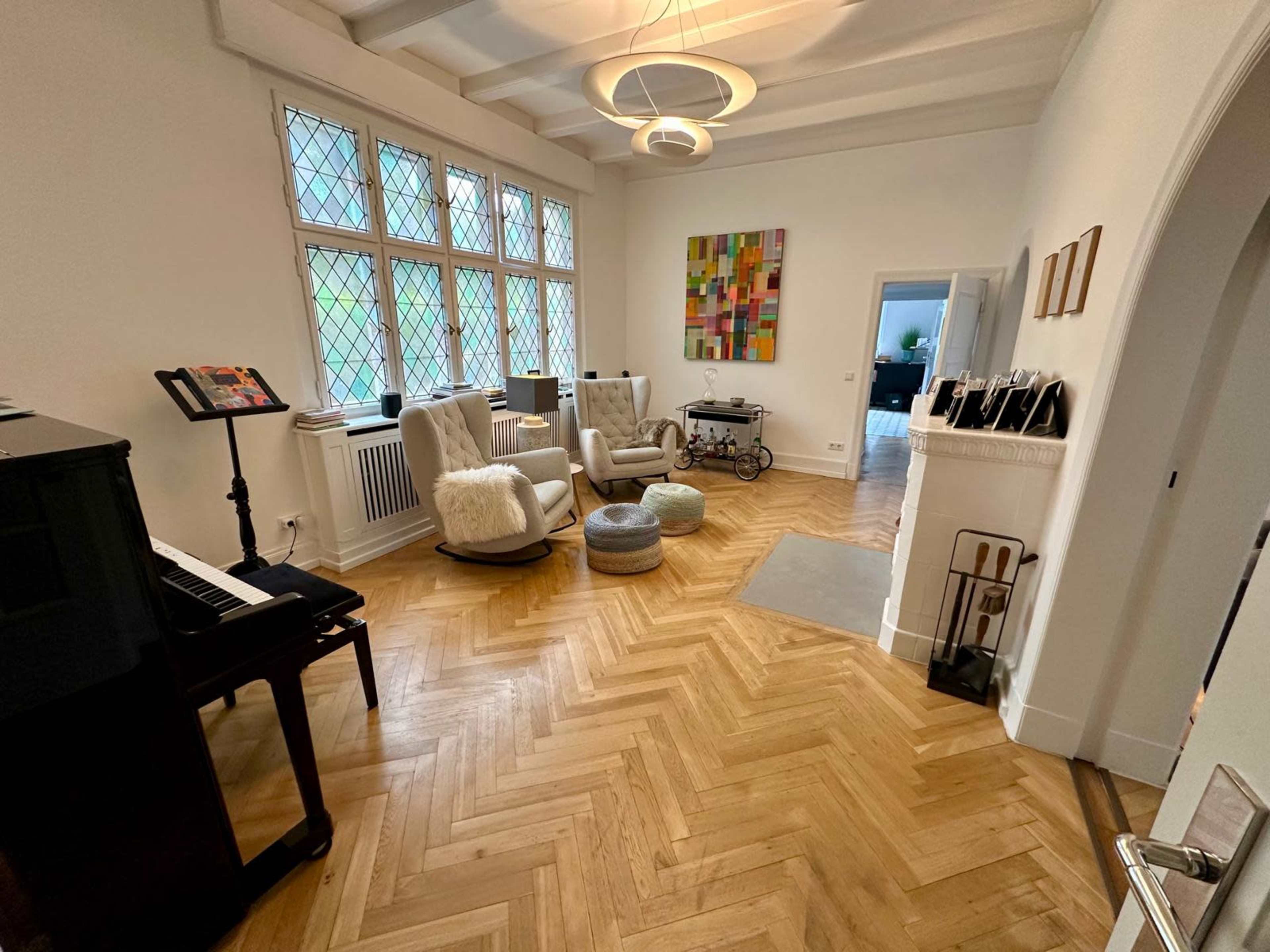 The image shows a spacious living room featuring two armchairs, a piano, a small cart, and a decorative painting on the wall.