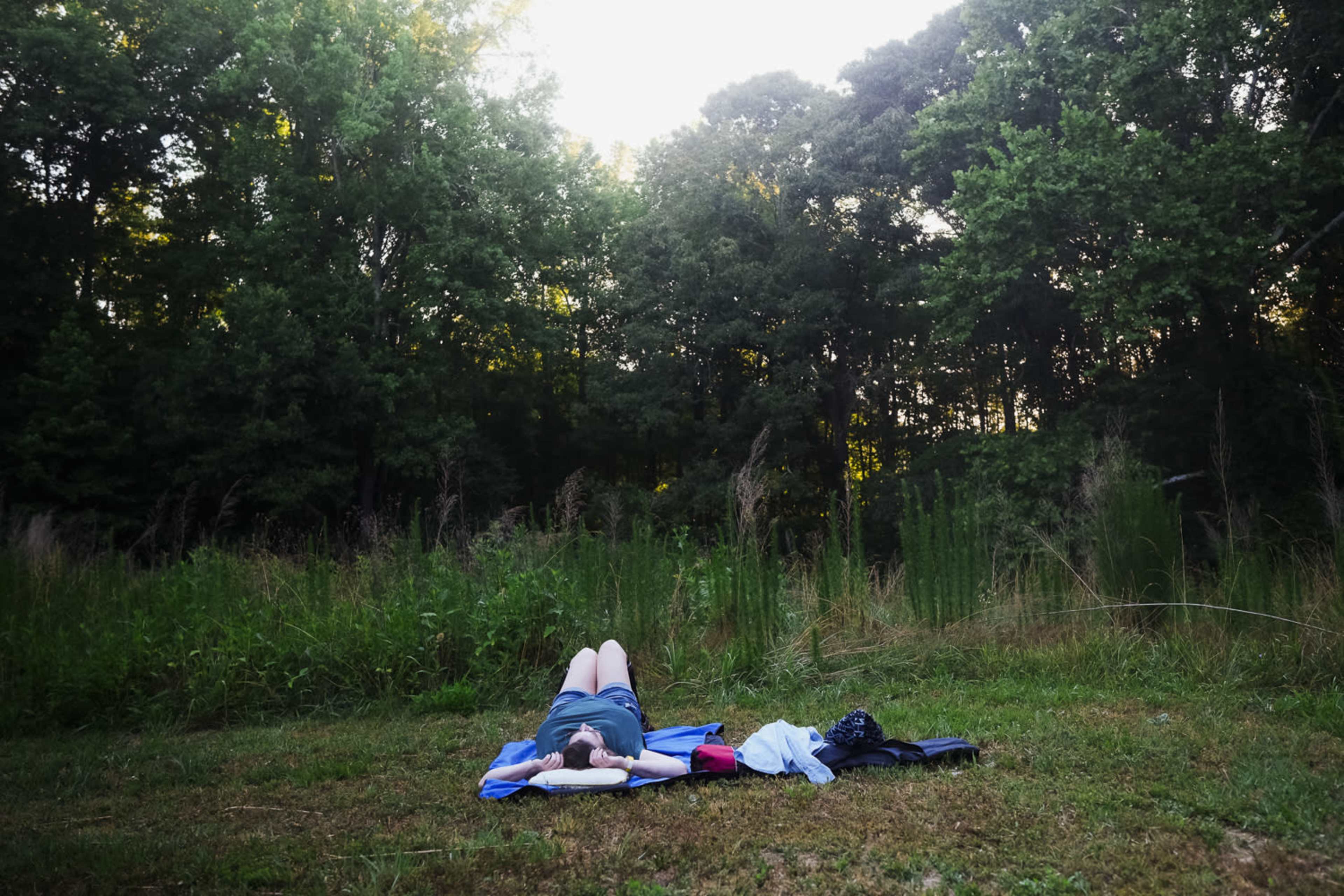 A person lies on a blue mat in a grassy area surrounded by tall trees and dense foliage.