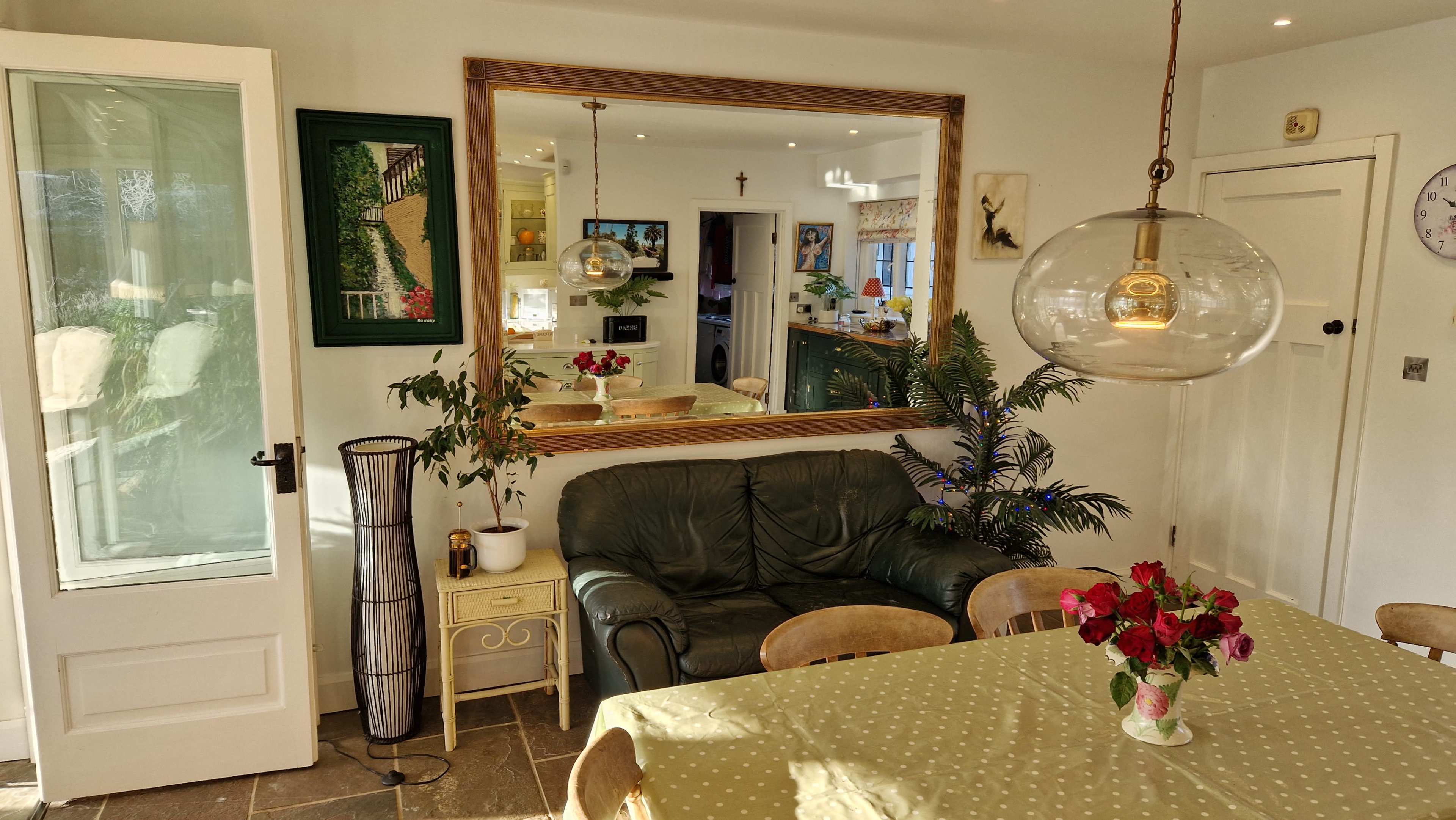 A spacious dining area features a large mirror, a green sofa, a wooden table with a polka dot tablecloth, and a vase of flowers, surrounded by various decorative elements.