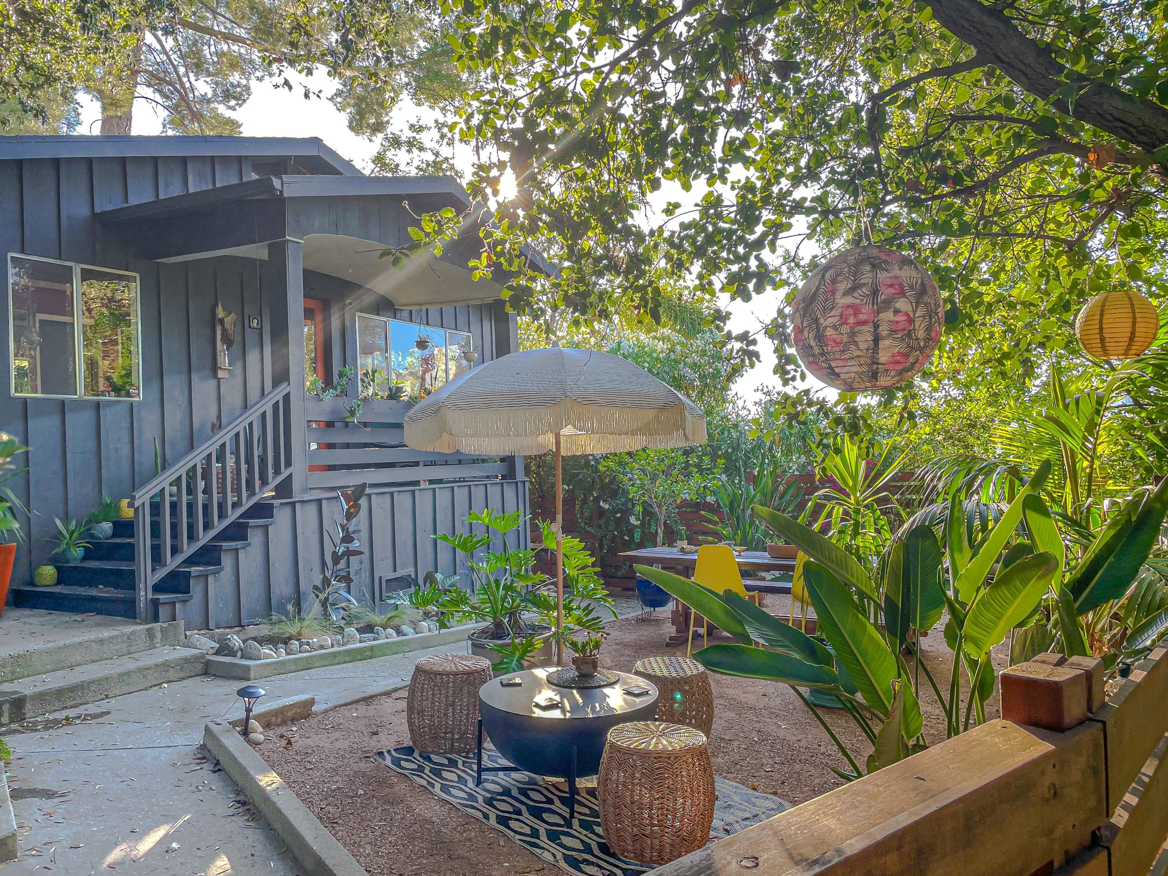 A cozy outdoor space features a seating area with a table and umbrella surrounded by lush plants and decorative lanterns.
