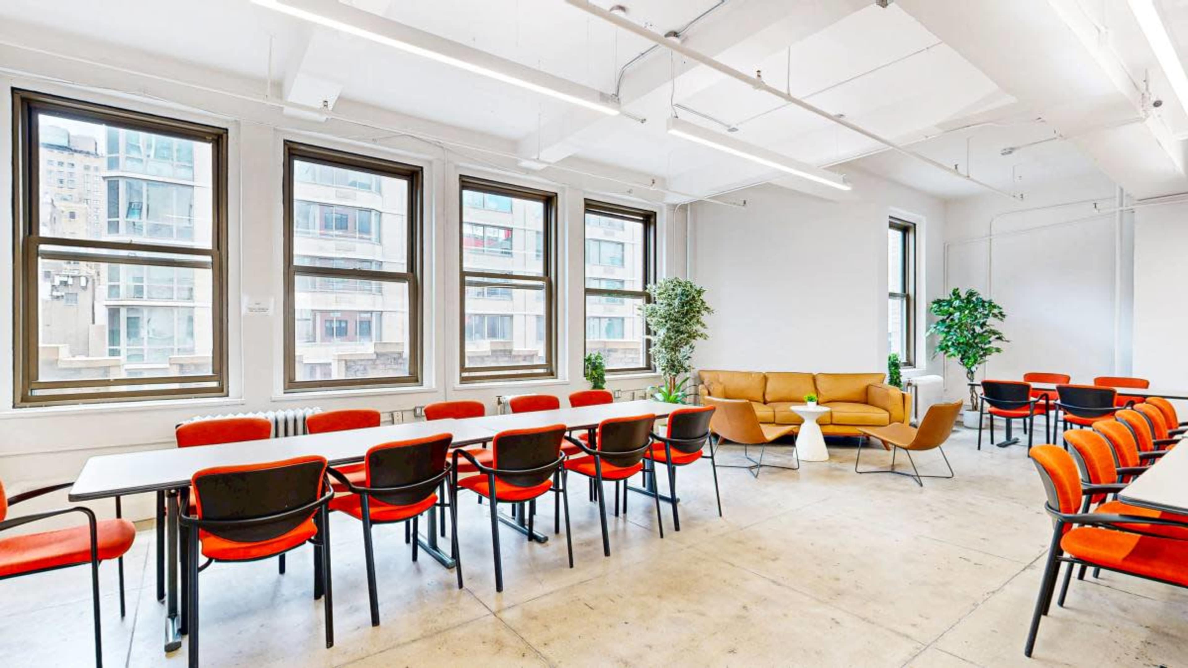 A bright, modern meeting room features a long table with chairs, a cozy sofa, and potted plants near large windows.