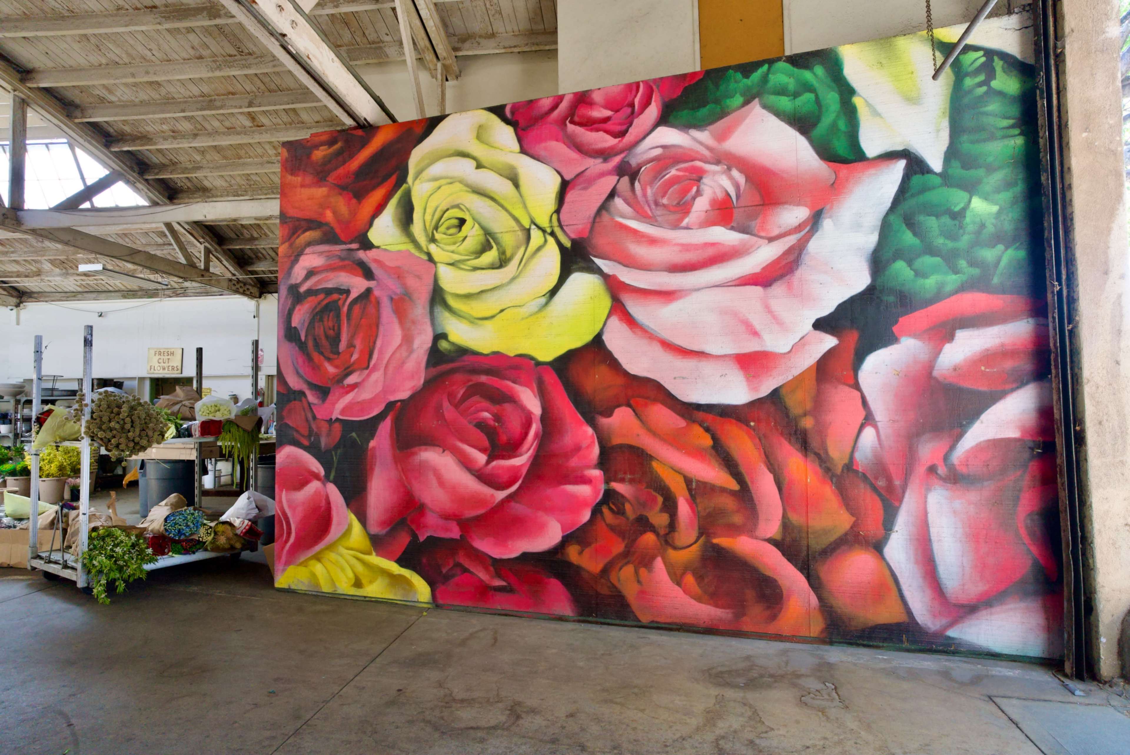 A large, vibrant mural of pink and yellow roses is painted on a wall, with a flower shop's display of plants and greenery visible in the background.