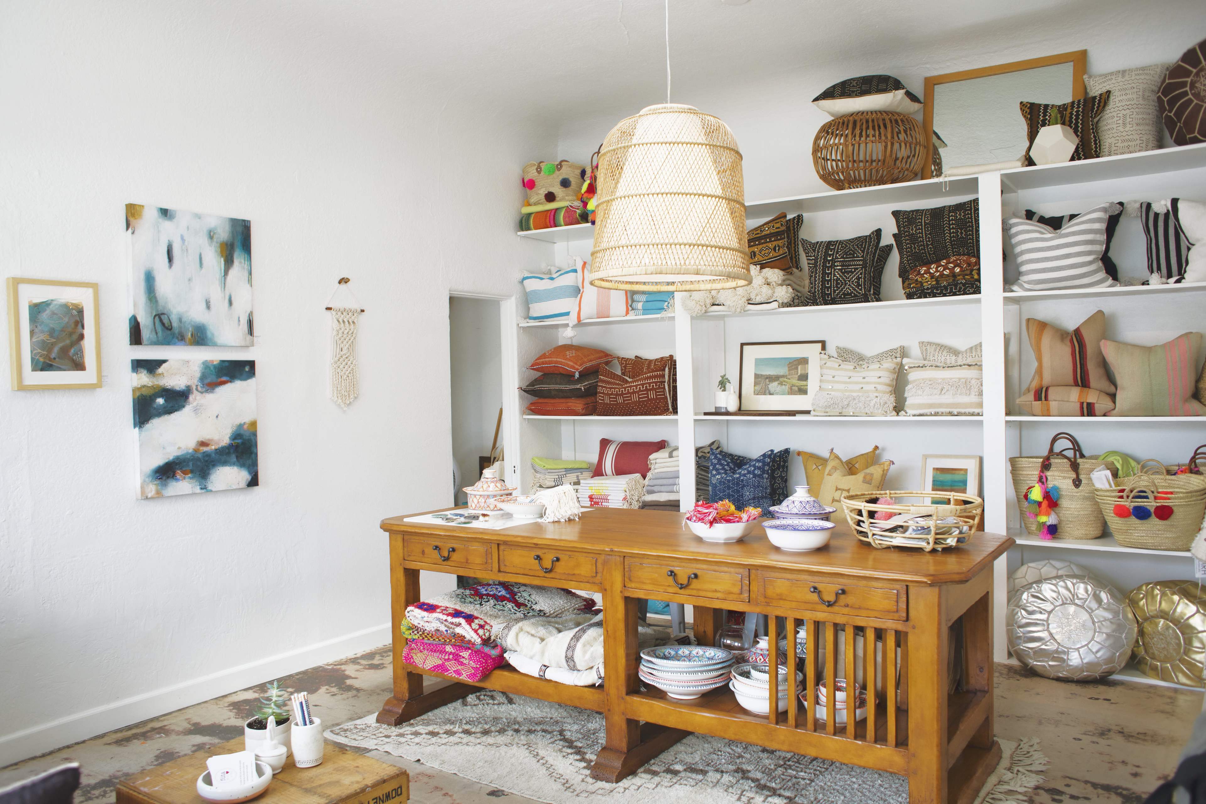 A bright, organized shop interior featuring a wooden table surrounded by shelves filled with decorative pillows, woven baskets, and colorful textiles.