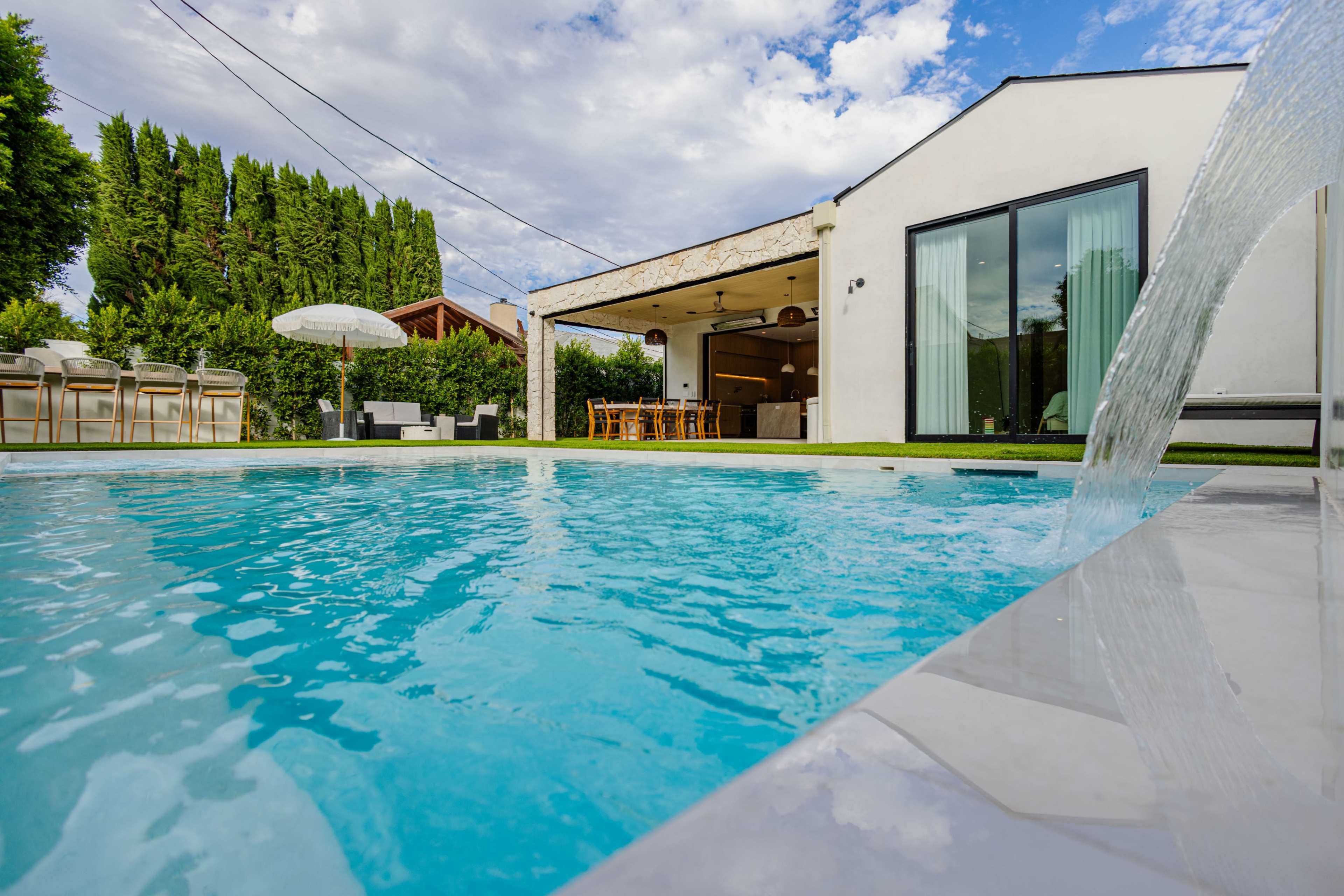 A clear blue swimming pool is in the foreground, reflecting the sky, with a modern home featuring large glass doors and outdoor seating visible in the background.