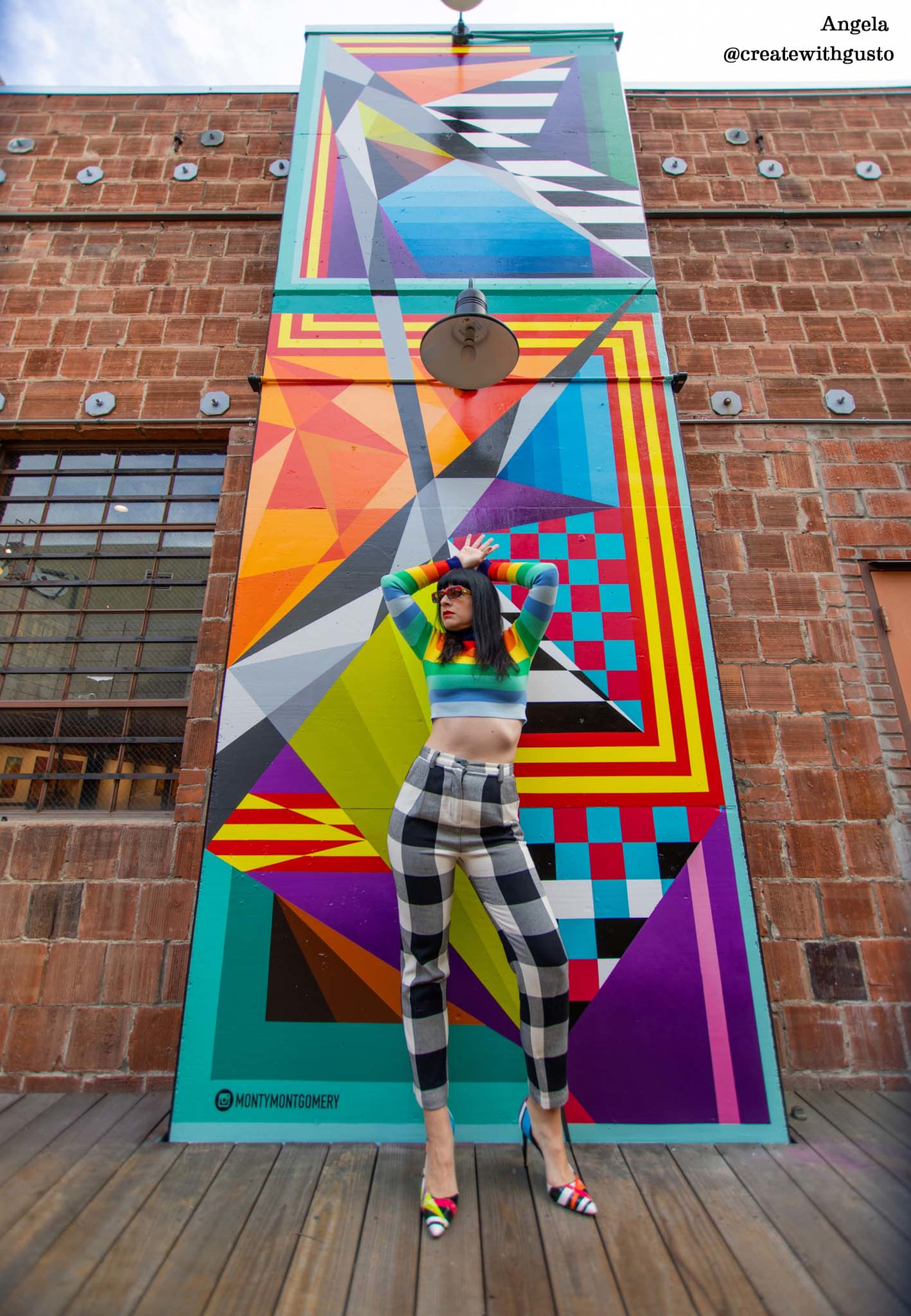 A woman poses in front of a colorful geometric mural, wearing a vibrant top and checkered pants.