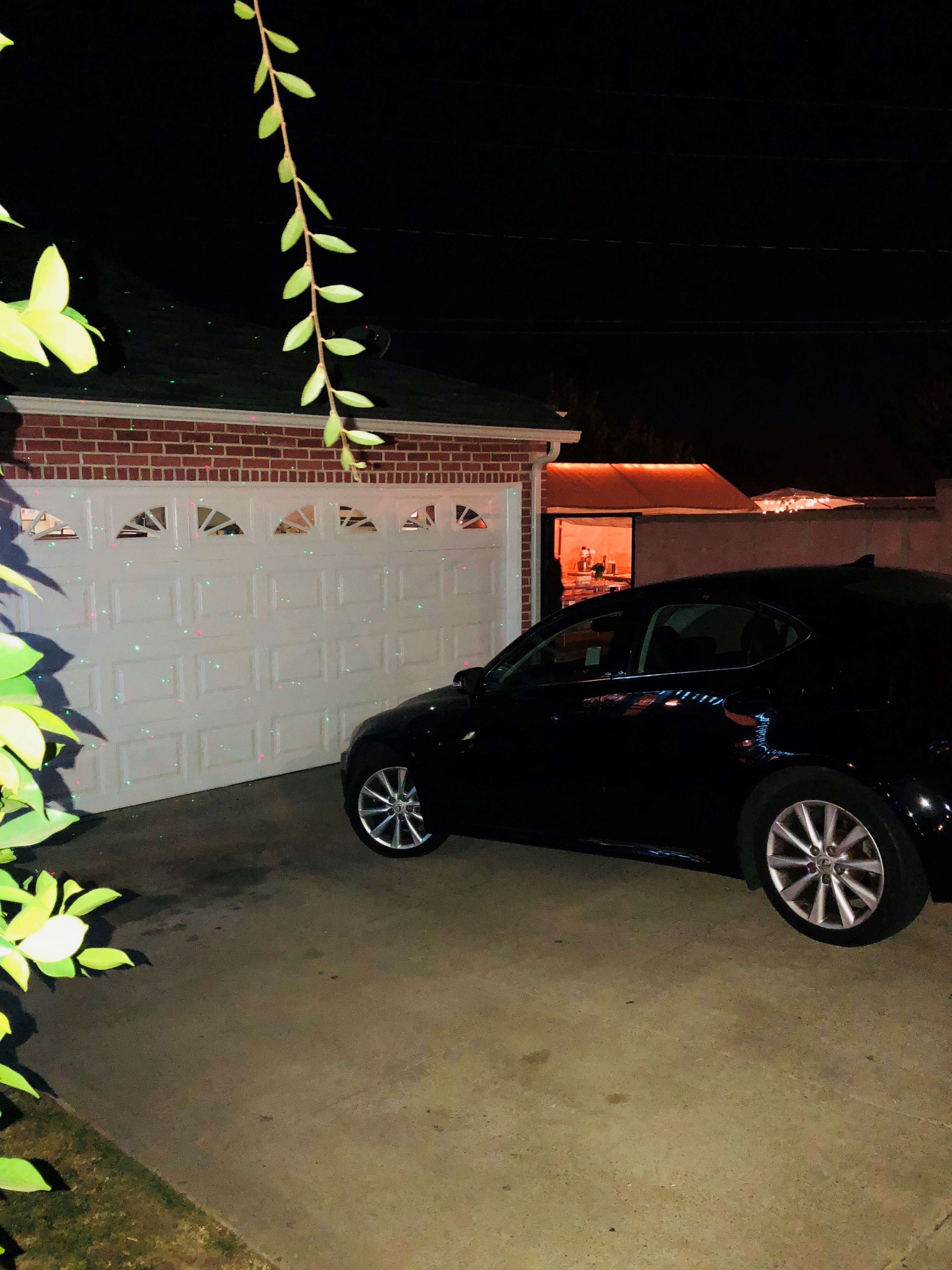 A black car is parked next to a white garage door at night, with a faint glow from lights in the background.
