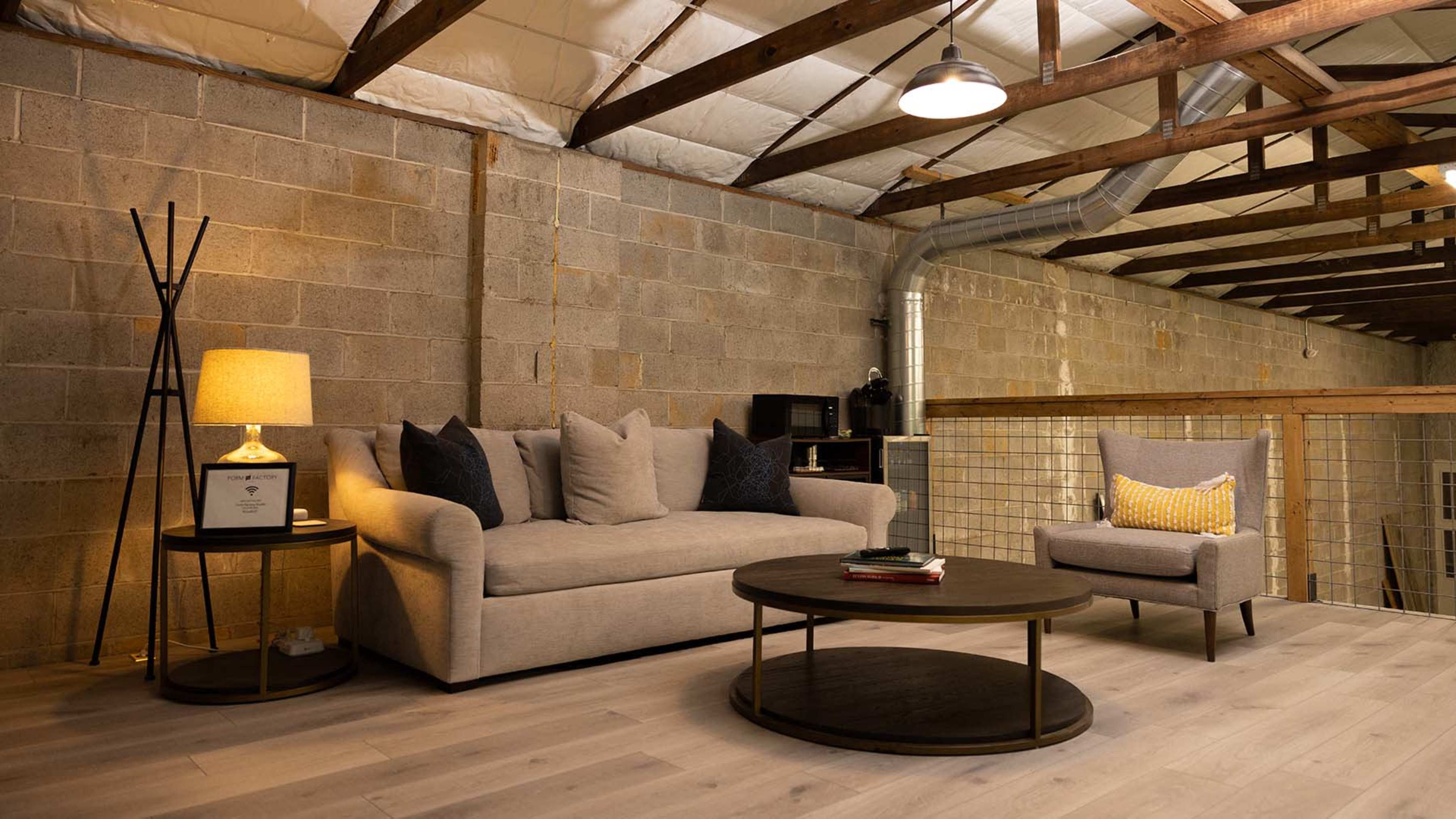 The image features a modern loft space with a beige sofa, a round coffee table, and a gray armchair, all set against a concrete wall and wooden beams.