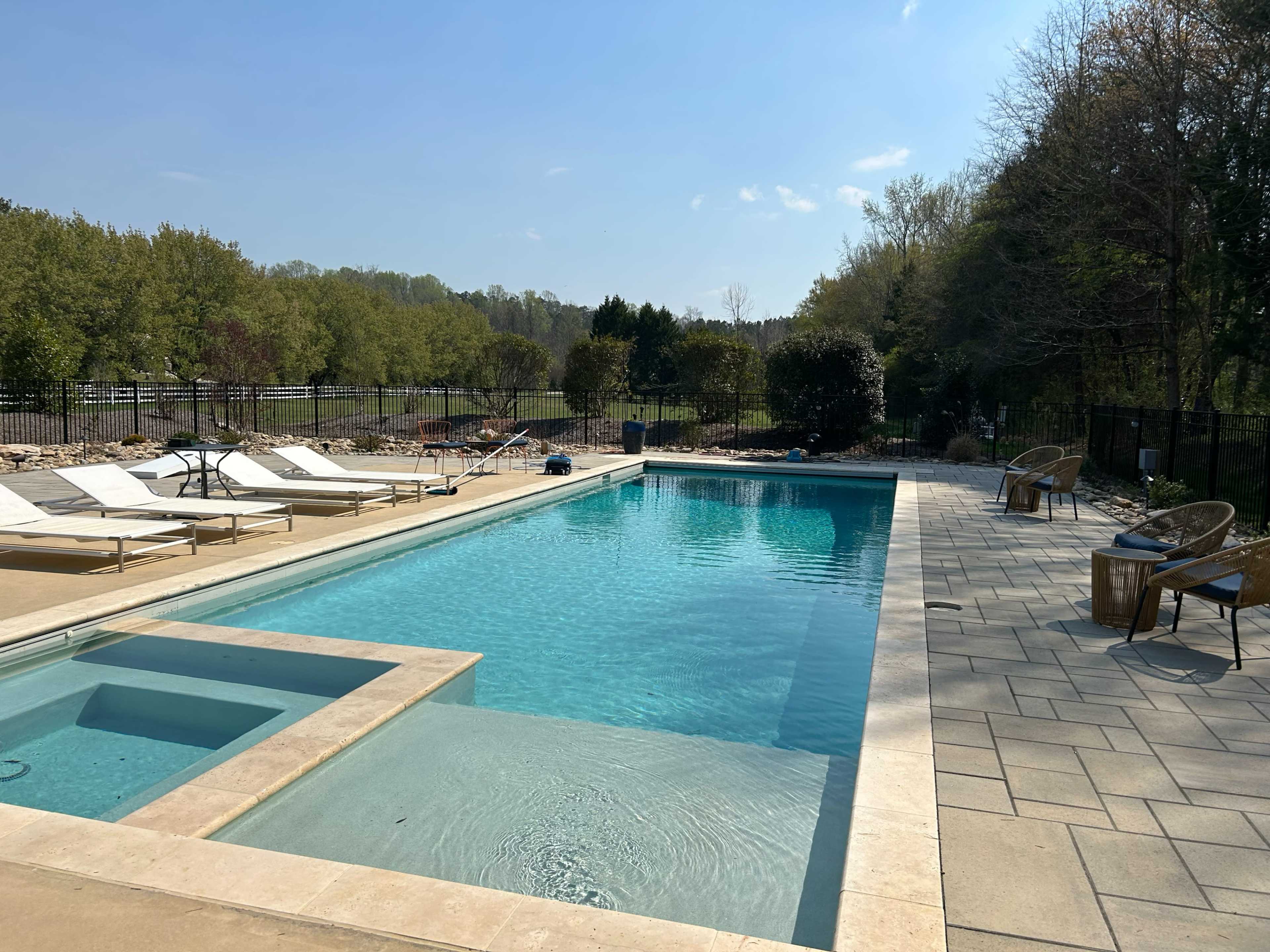 The image shows a swimming pool surrounded by lounge chairs, with a landscaped area and trees in the background.