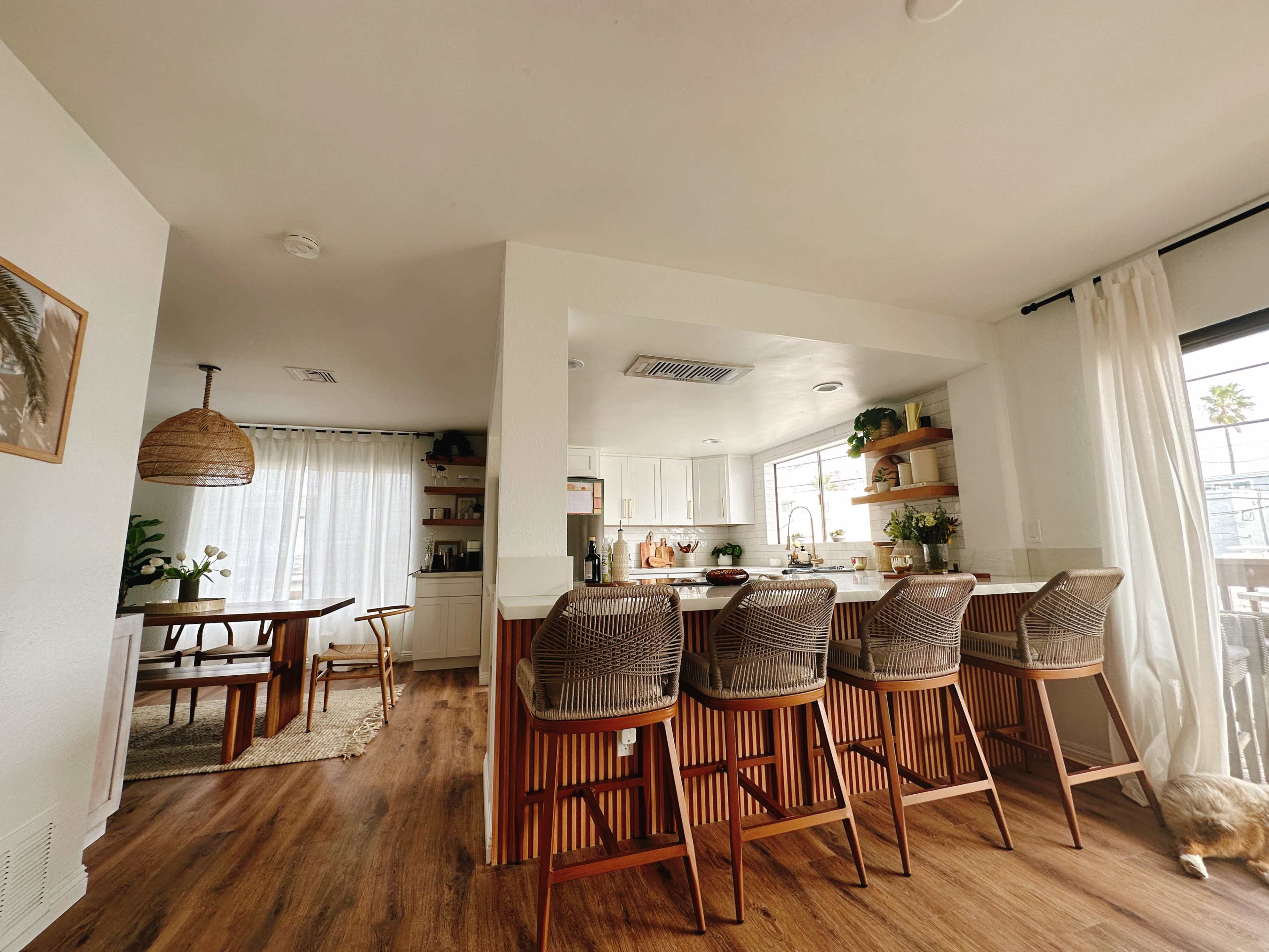 A modern kitchen with bar stools, a dining area, and large windows allowing natural light to enter.