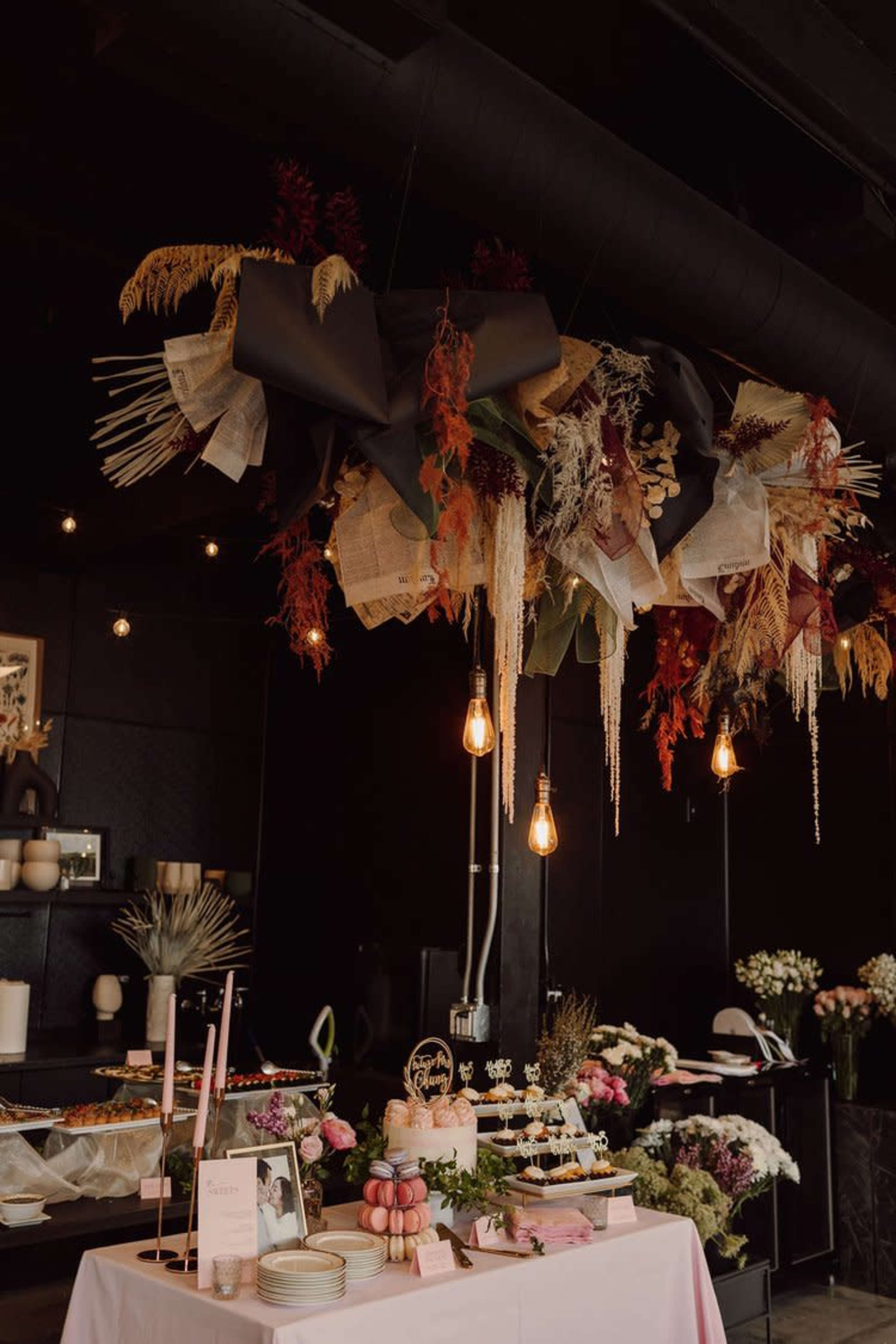 A decorated table with a cake and pastries is set beneath a floral and textured ceiling installation in a dimly lit room.