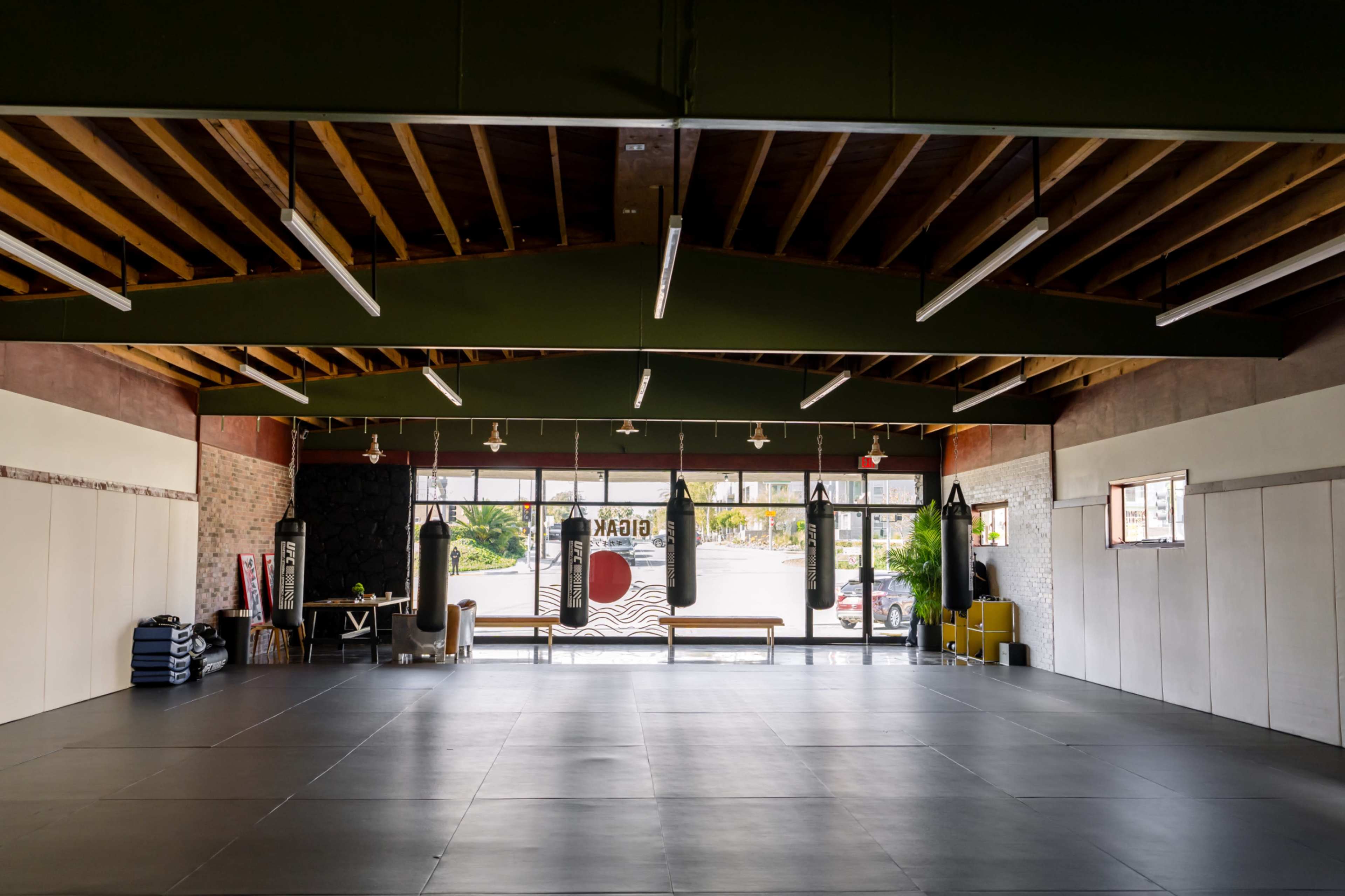 The image shows a spacious martial arts studio with padded flooring, large windows, and several punching bags hanging from the ceiling.