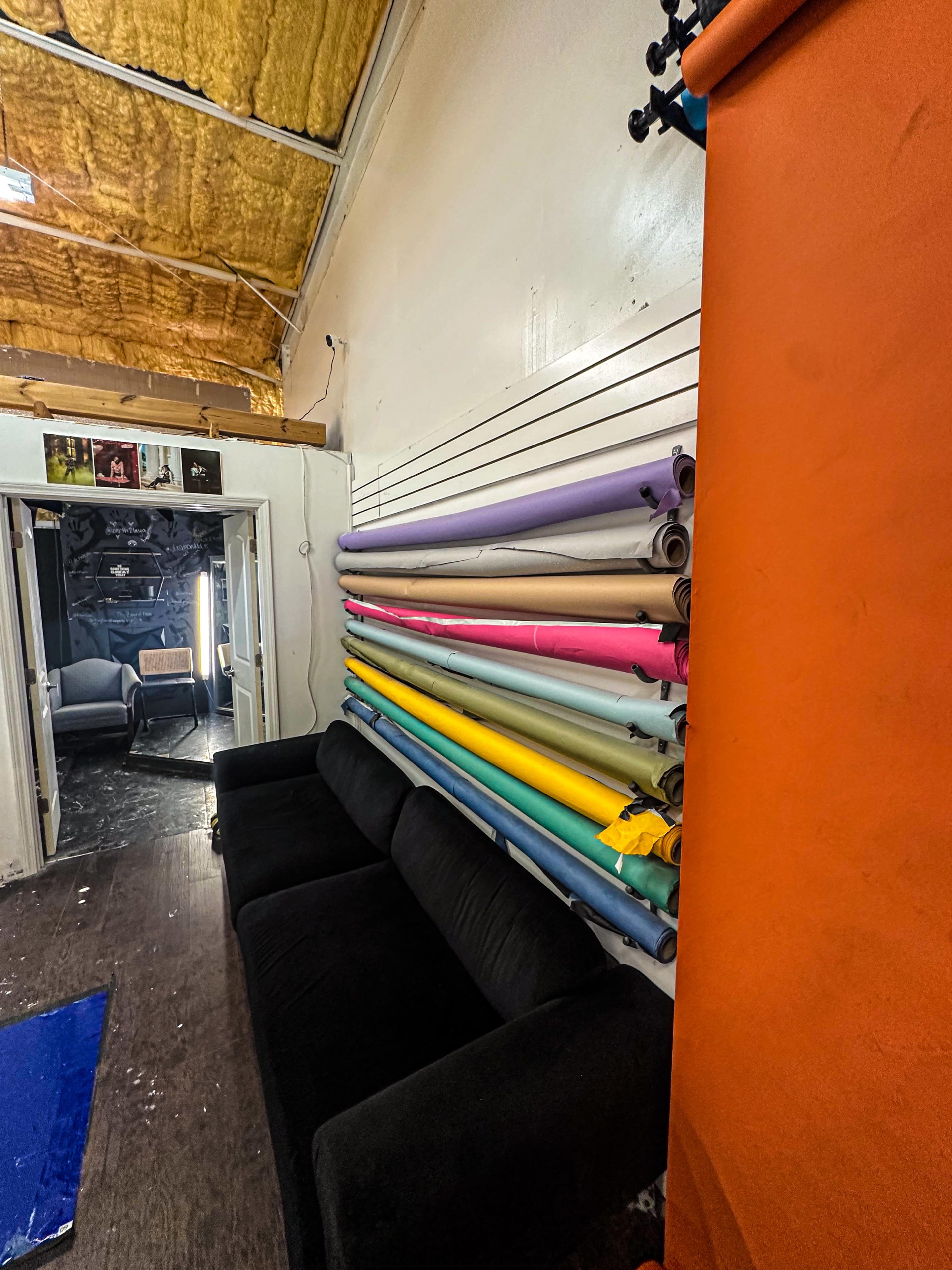 The image shows a black couch against a wall with colorful rolls of backdrop paper arranged horizontally above it, in a room with a textured ceiling.