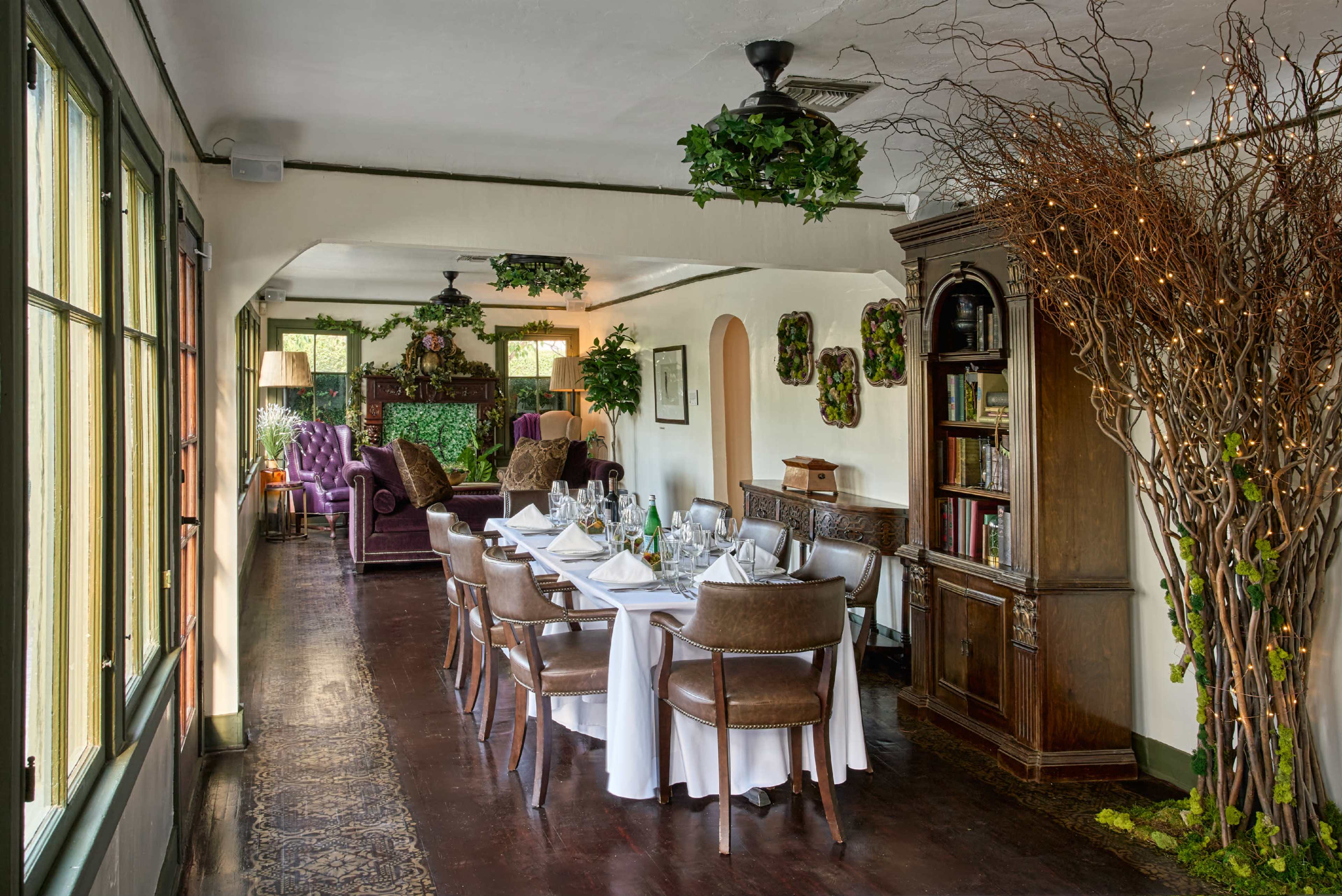 A well-decorated dining area with a long table set for a meal, surrounded by plants and wooden furniture.