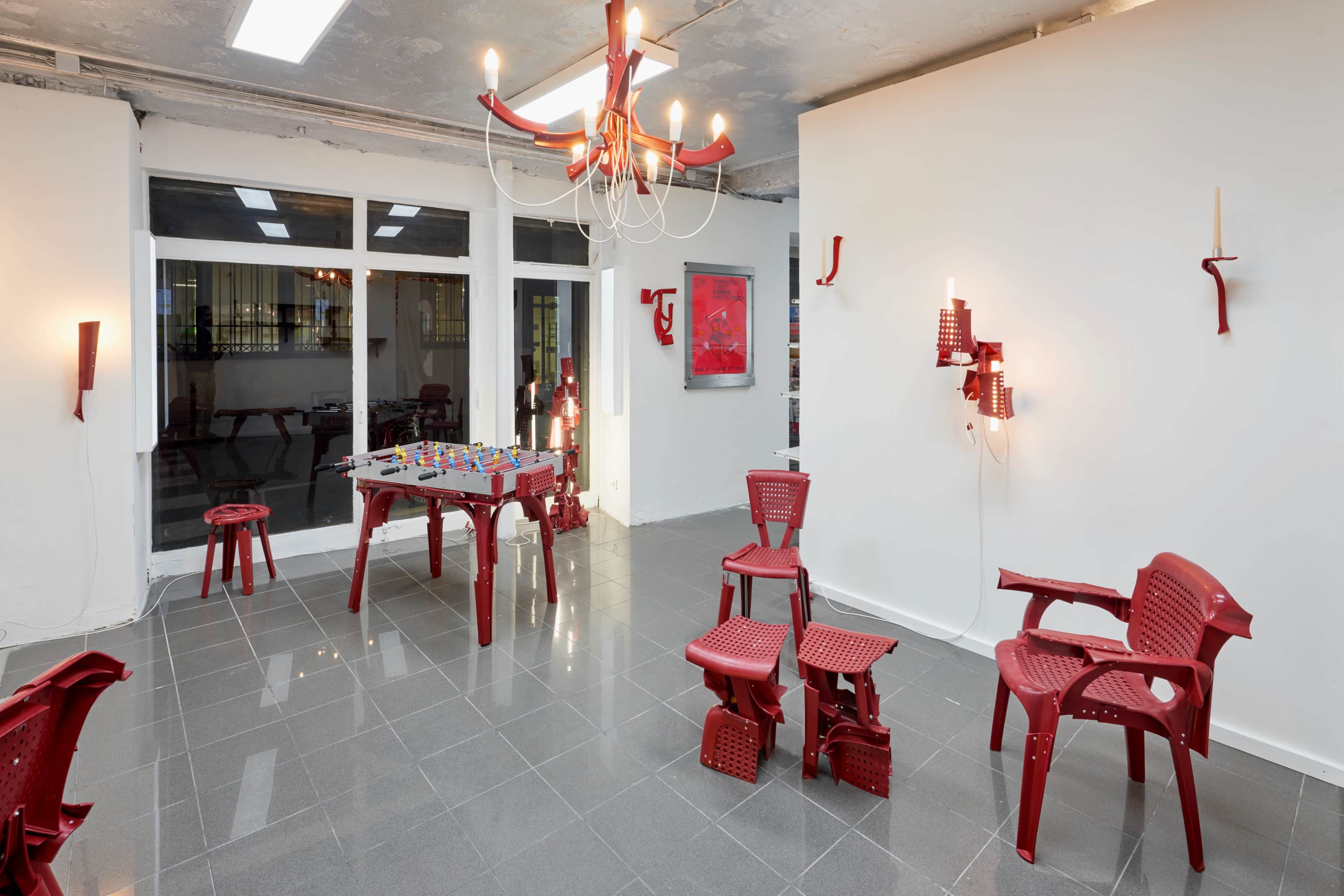 The image shows an art gallery featuring a variety of red plastic chairs, a game table, and wall-mounted sculptures, all arranged in a modern space with gray tiled flooring.