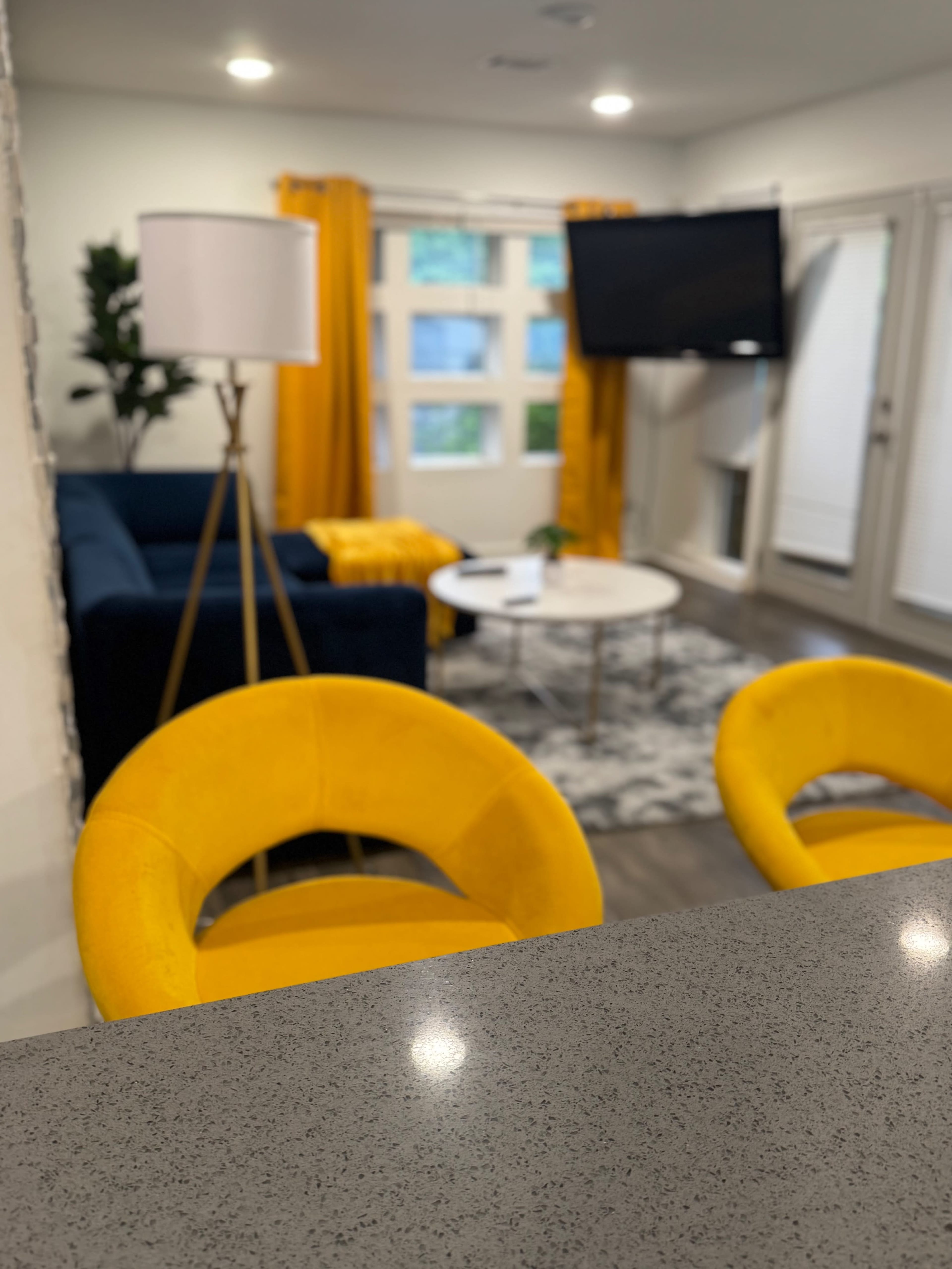 A modern living room features a blue sofa, a round coffee table, and yellow curtains, with two yellow chairs in the foreground.