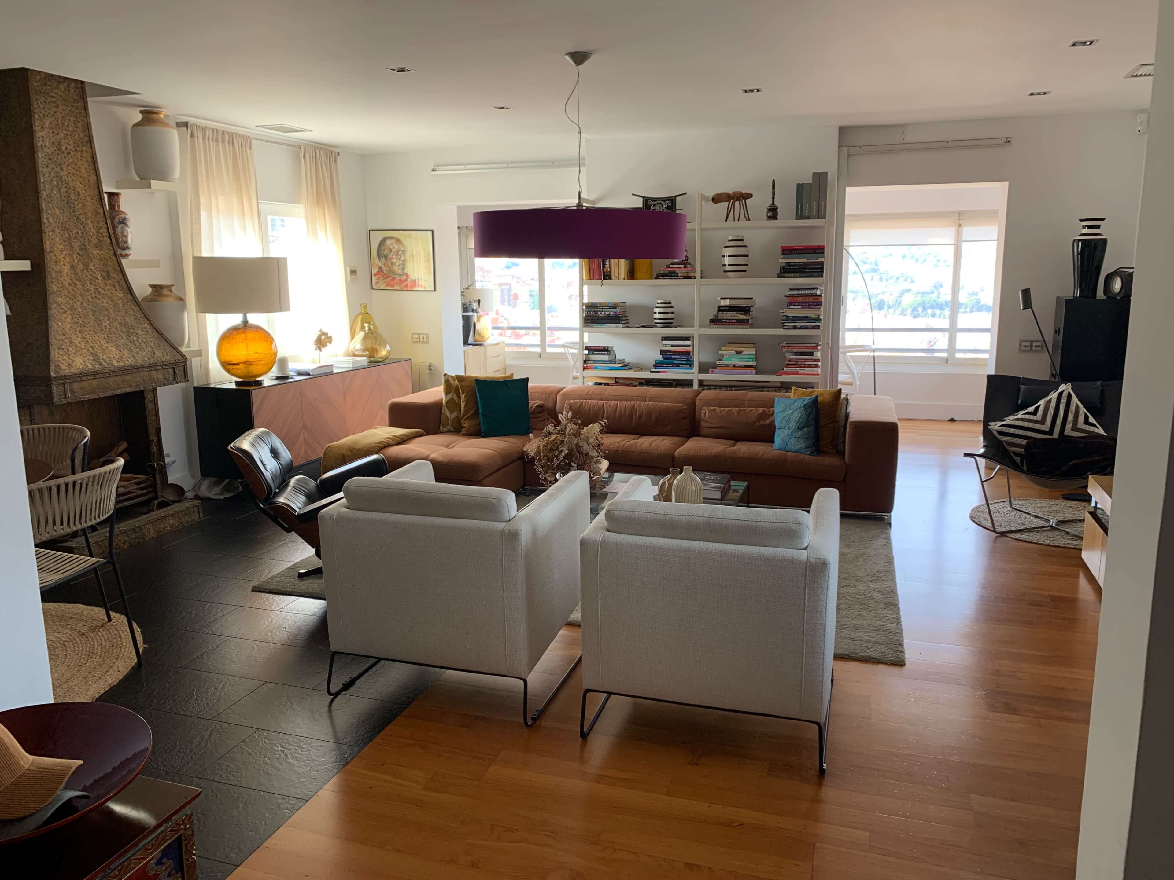A modern living room featuring a brown sofa, two white chairs, a coffee table, and shelves filled with books, with large windows allowing natural light.