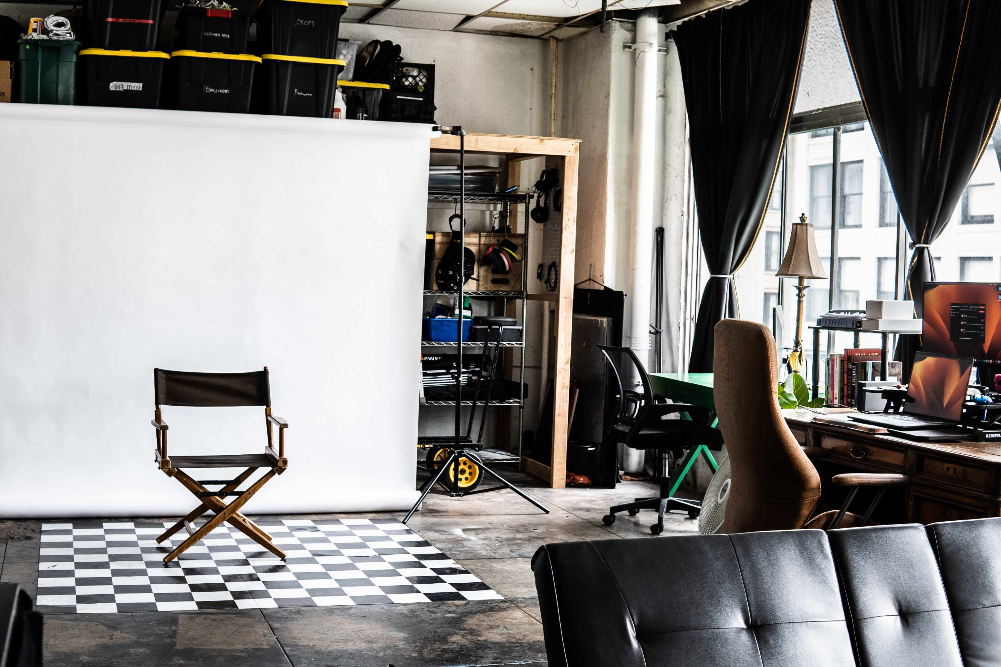 A studio space features a folding chair in front of a white backdrop, a checkered floor, and various equipment organized on shelves.
