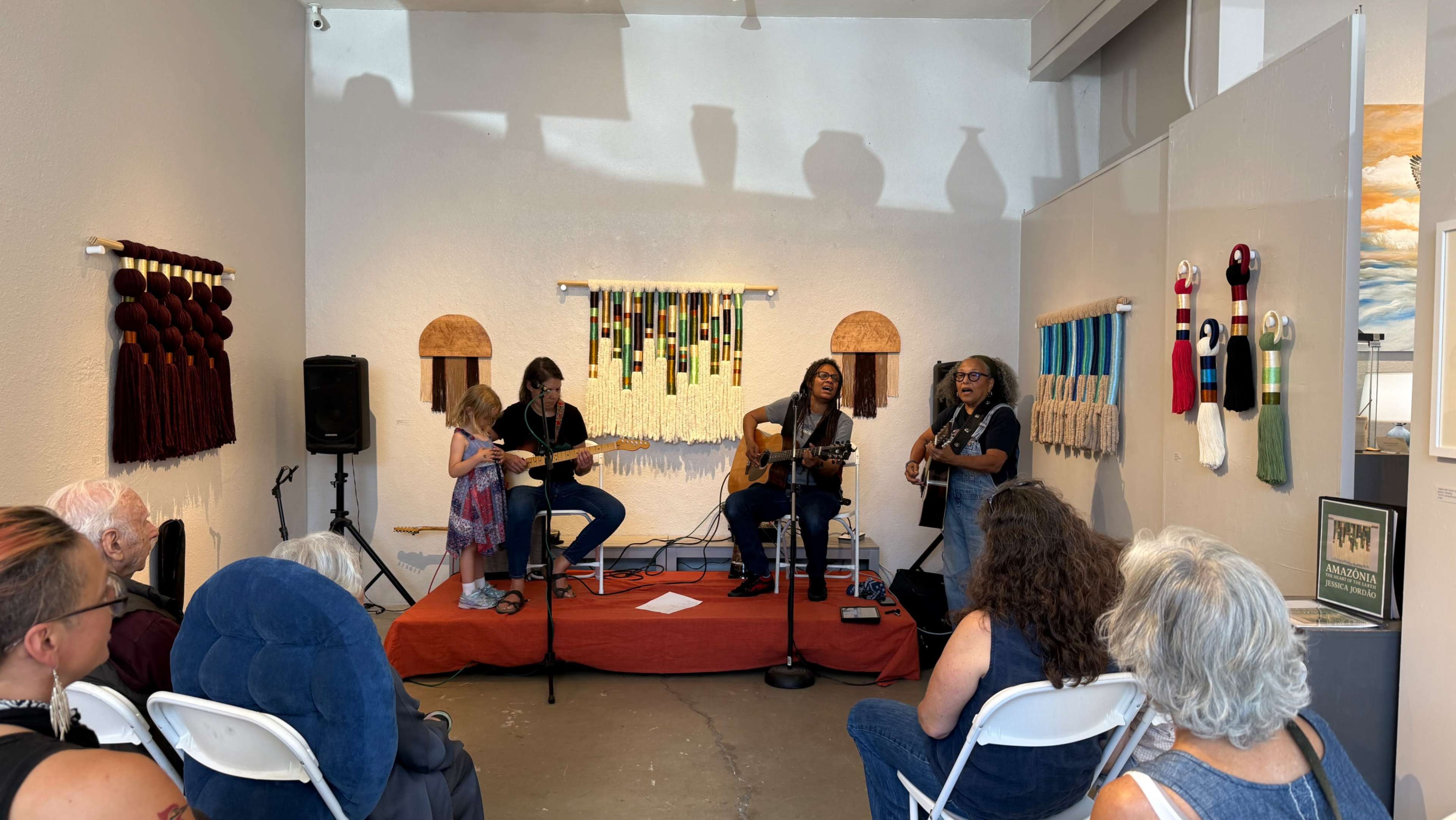 Three musicians perform on a red rug in an art space, while an audience watches from white chairs.