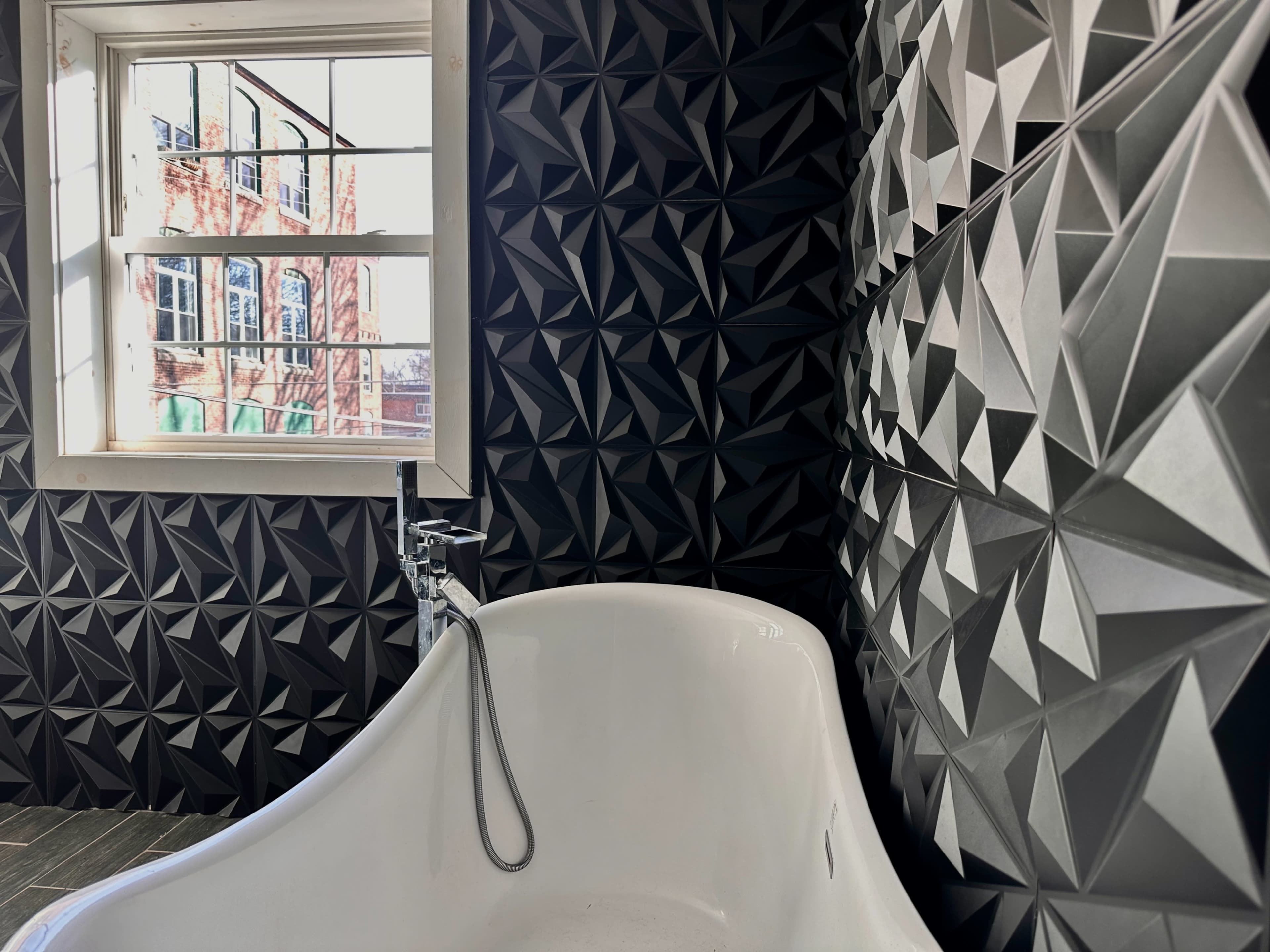A bathroom with a white bathtub and textured black and gray geometric wall tiles, bathed in natural light from a nearby window.