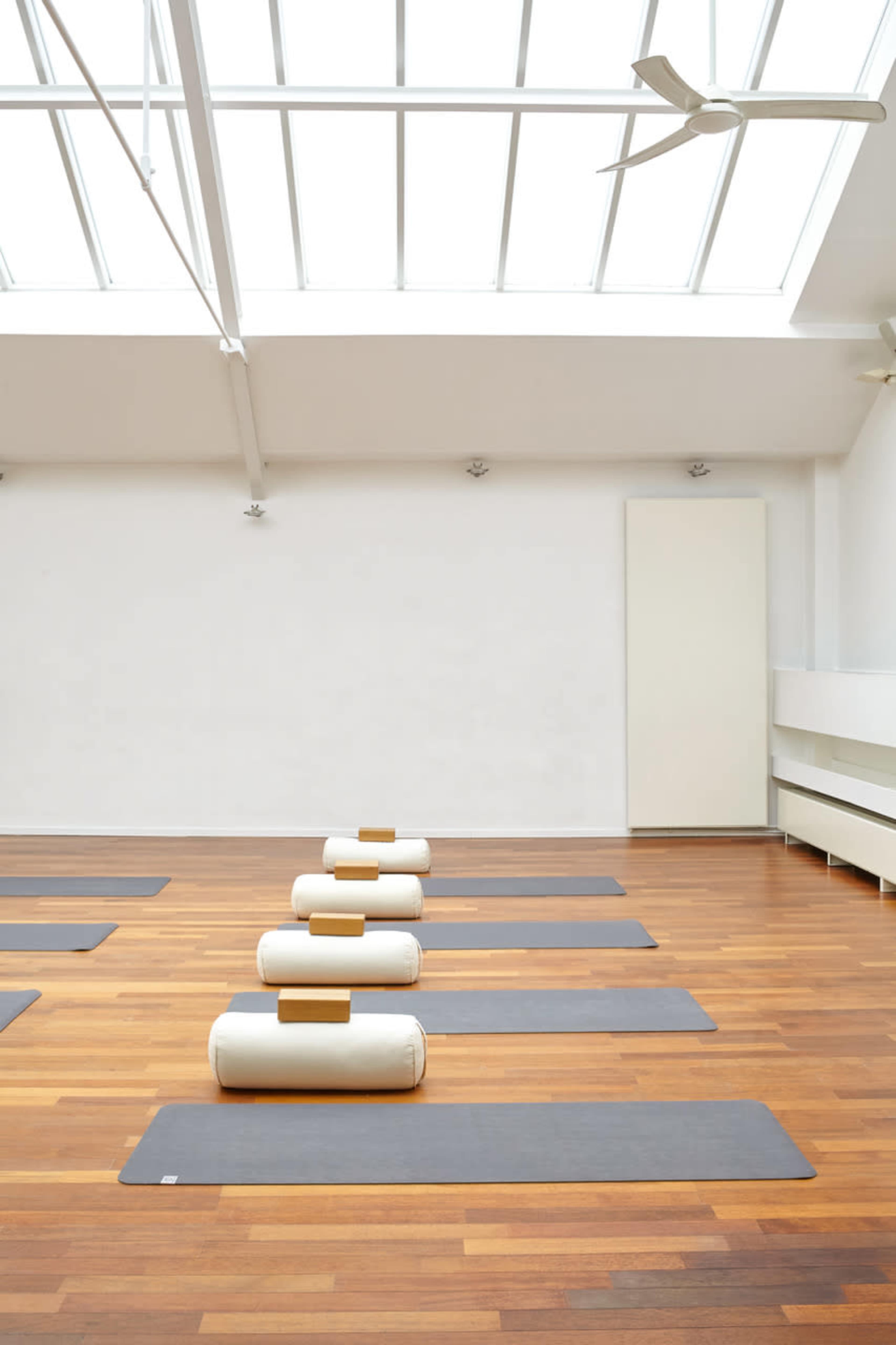 A spacious yoga studio with wooden floors, light gray yoga mats, and white bolster pillows arranged in a neat row under a skylighted ceiling.