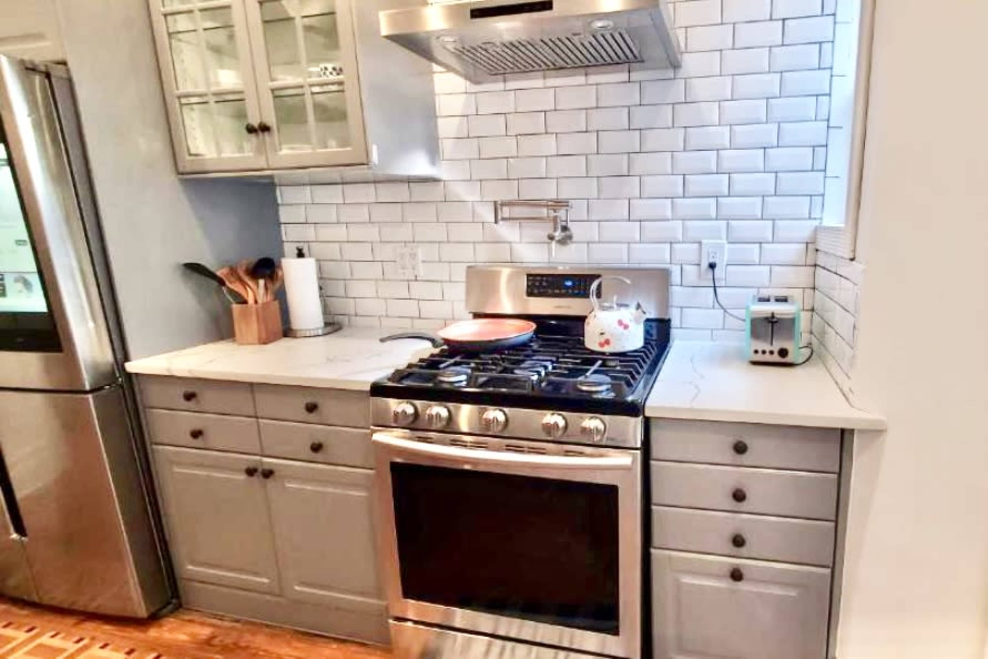 The image shows a modern kitchen featuring a stainless steel gas stove beneath a metal hood, grey cabinetry, white subway tile backsplash, and a countertop with kitchen utensils and a kettle.