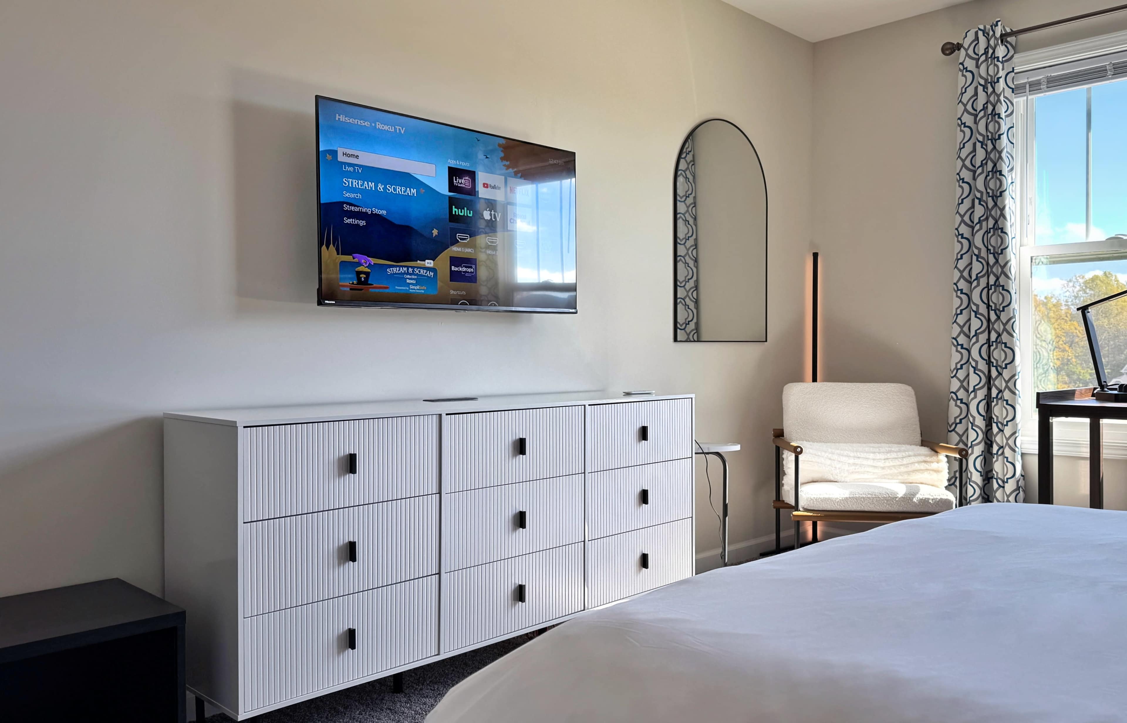 The image shows a modern bedroom featuring a white dresser, a mounted TV on the wall, a mirror, a chair, and a window with curtains.