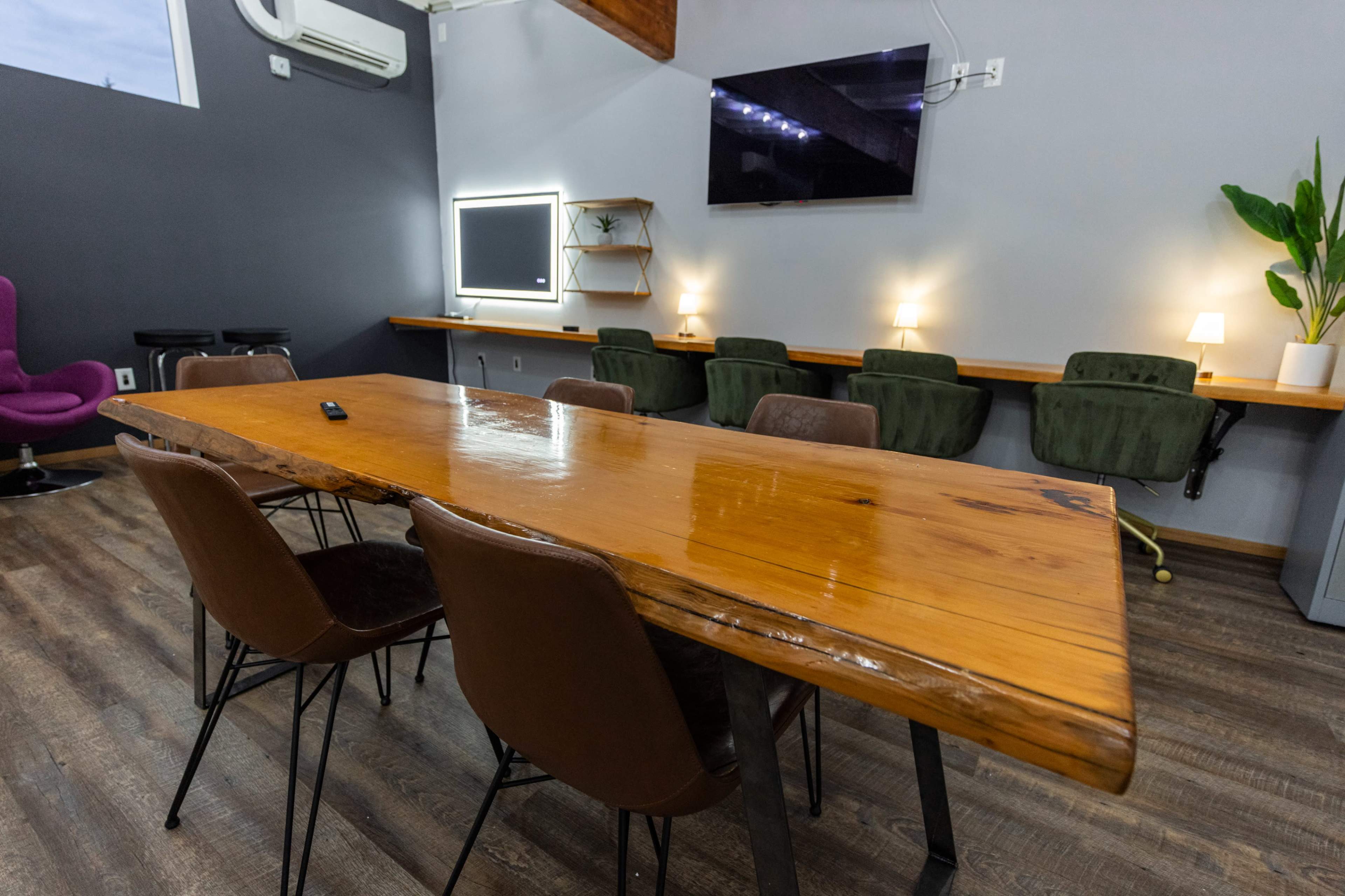 A spacious meeting room featuring a large wooden table surrounded by chairs, with wall-mounted screens and plants in the background.