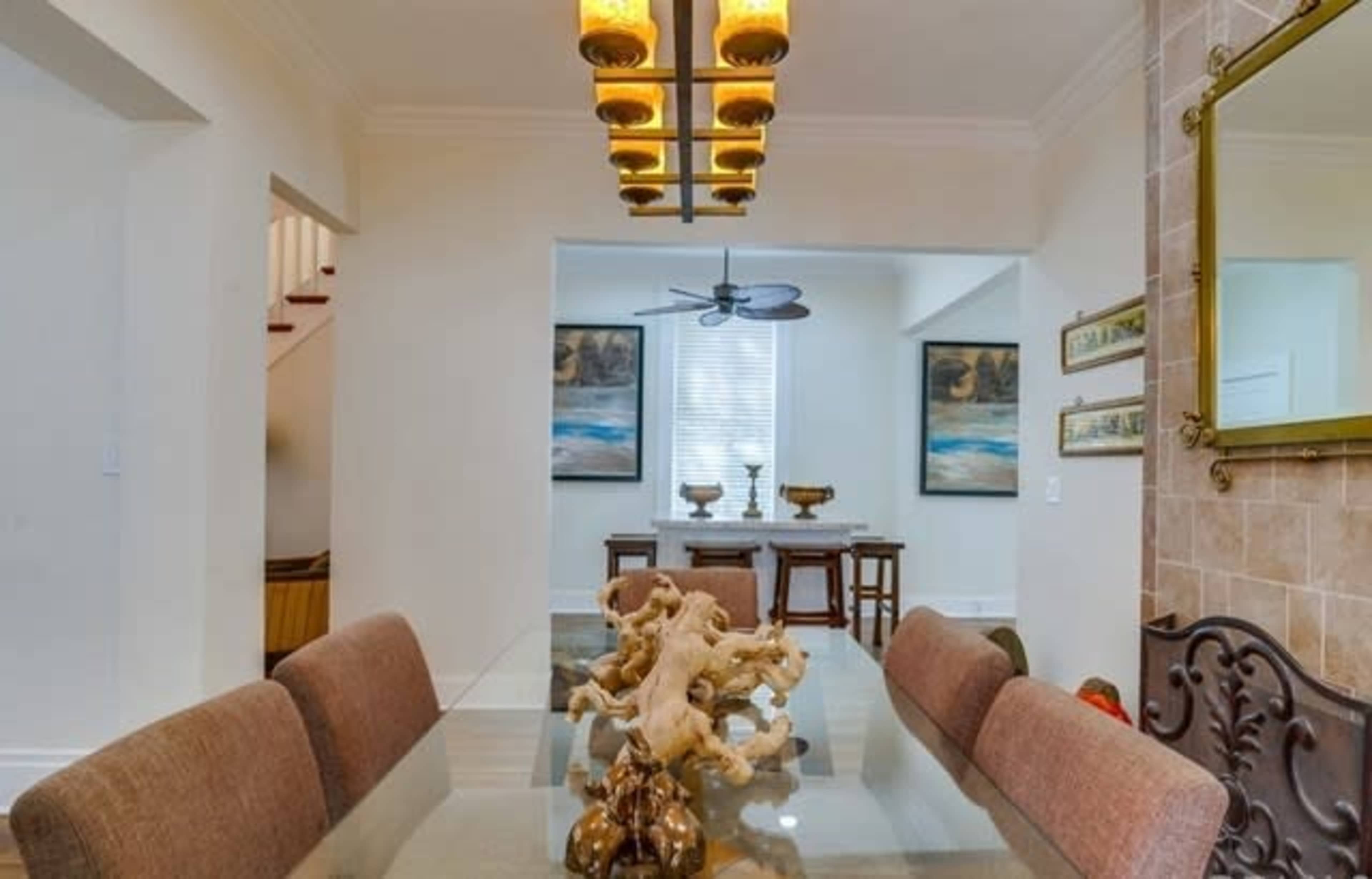 A modern dining area features a long glass table with decorative elements, illuminated by a stylish chandelier, with a view of an adjacent room.