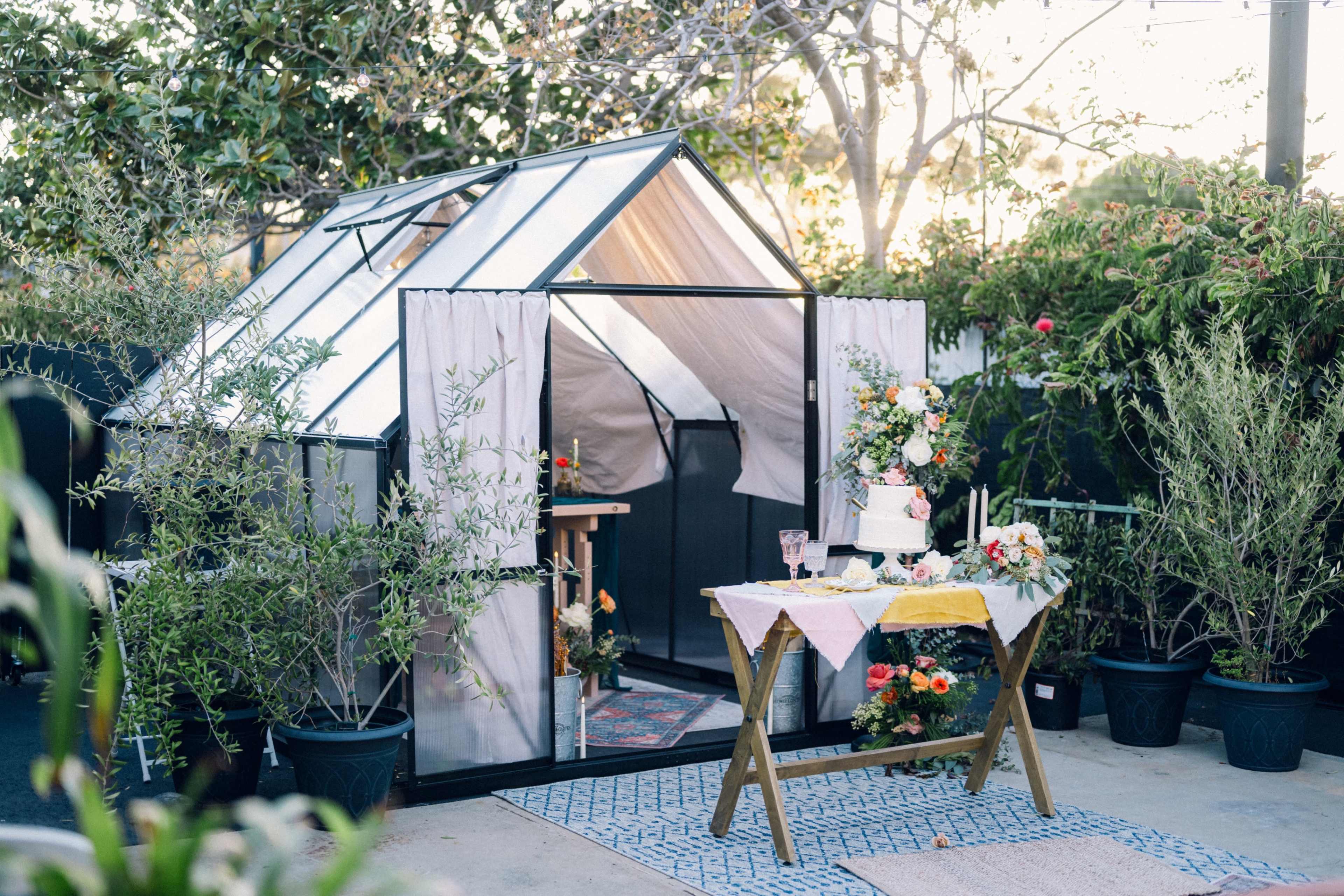 A glass greenhouse is surrounded by potted plants, with a table adorned with a cake and floral arrangements set nearby.
