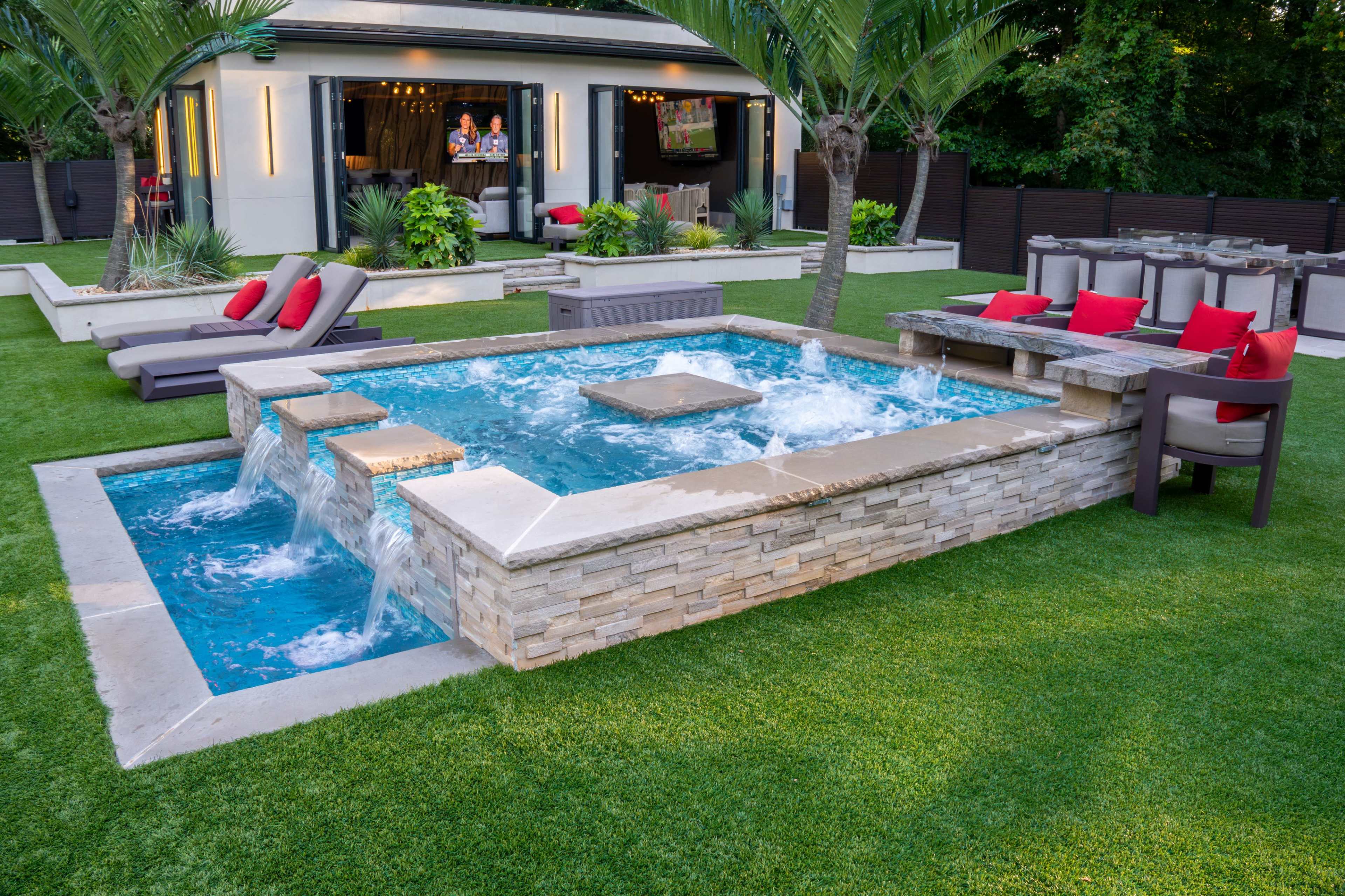 The image shows a backyard pool area featuring a raised spa with water cascading over stone edges, surrounded by lounge chairs and a patio with outdoor seating.