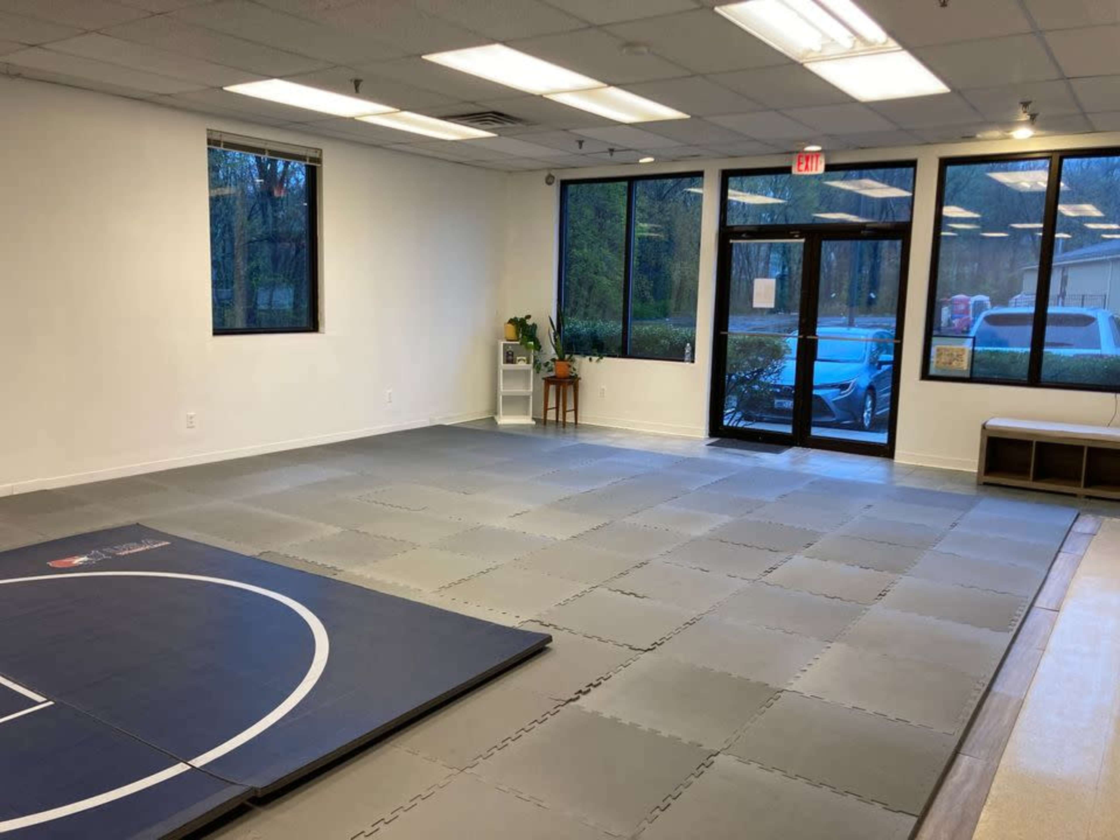 An empty training room with a mat in one corner and large windows allowing natural light to enter.