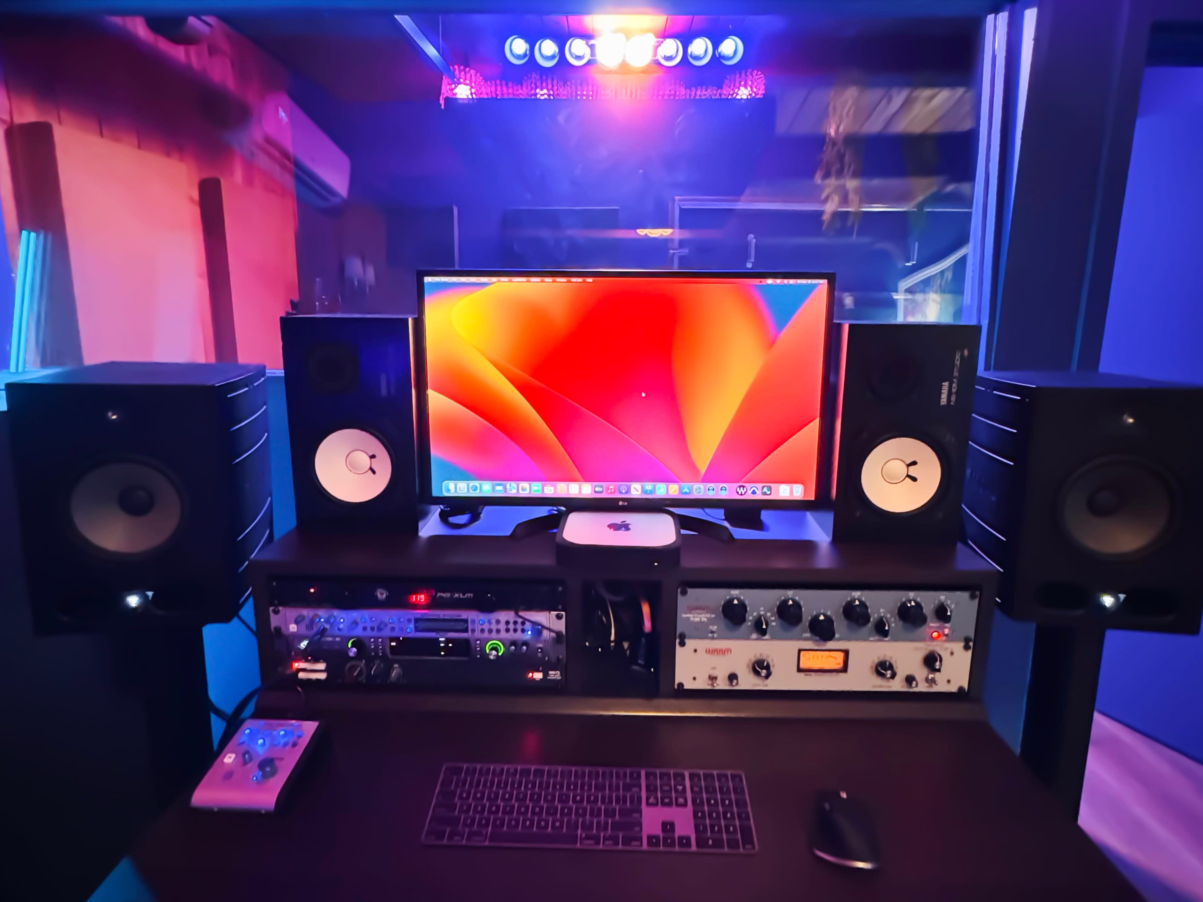 A music production studio with a computer monitor, speakers, audio equipment, and a keyboard on a dark desk.