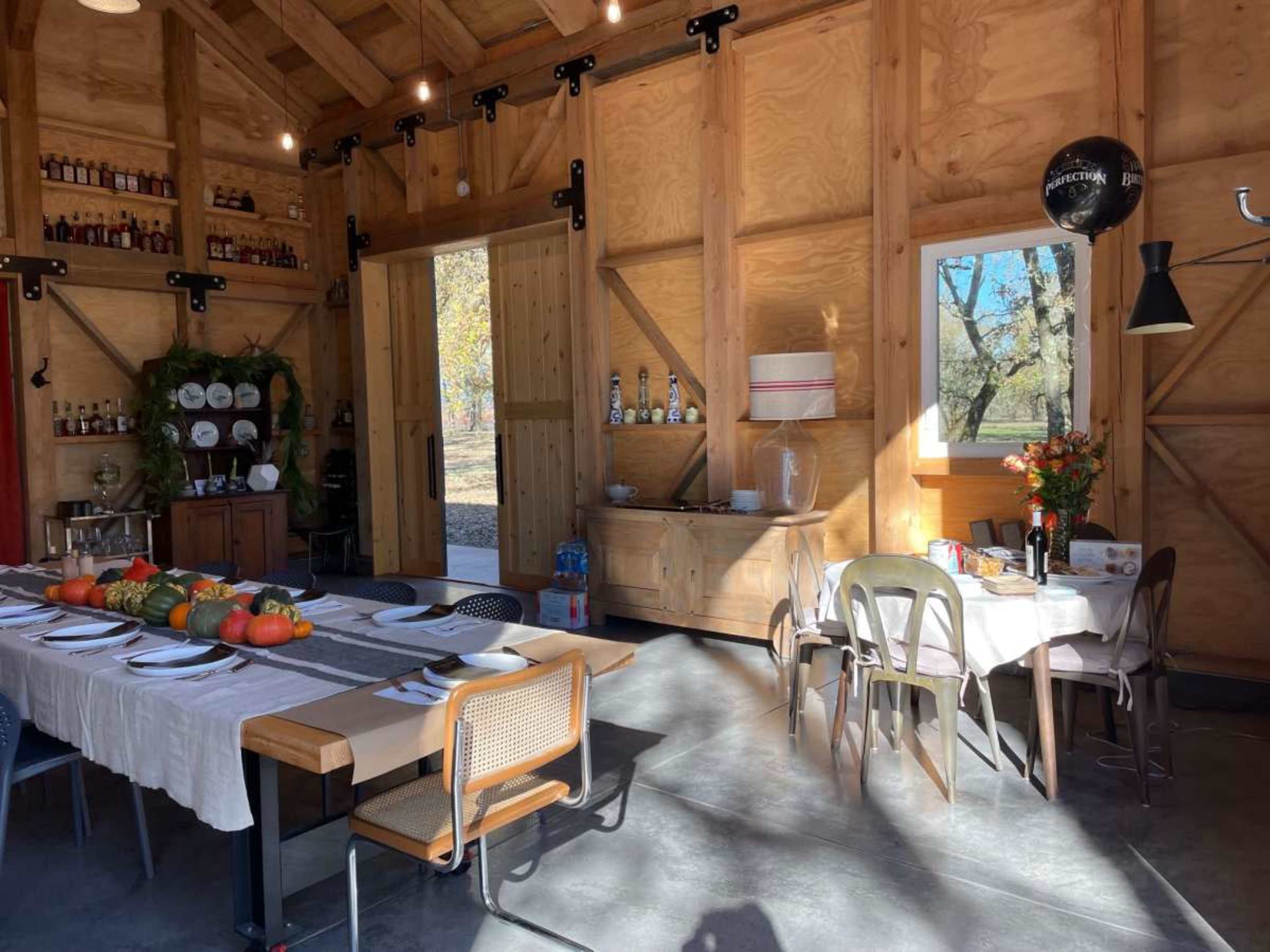 The image shows an interior of a wooden building featuring a dining area with a long table set with plates and pumpkins, alongside a bar area with shelves of bottles.