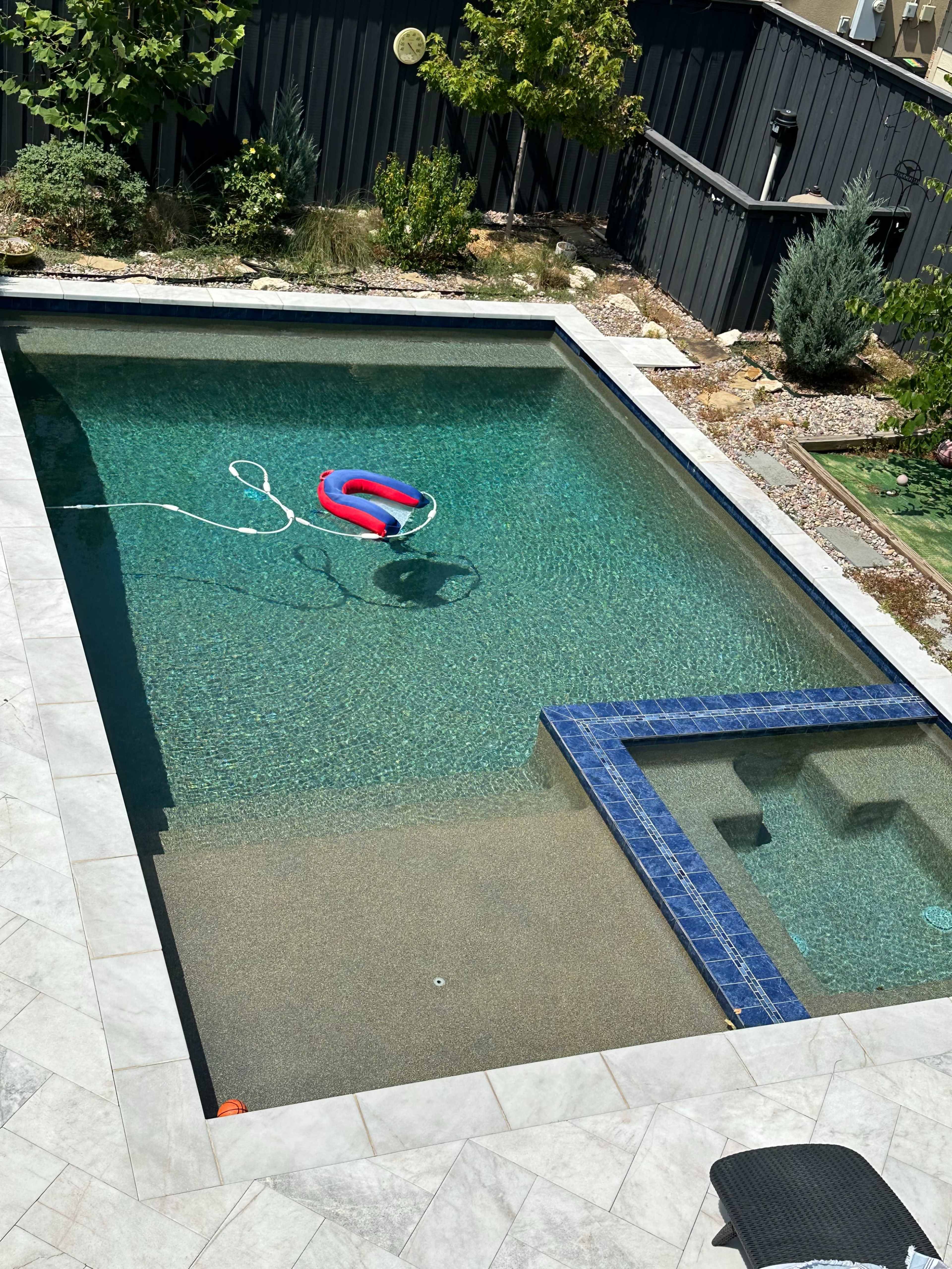 A clear swimming pool features a colorful float and a spa area surrounded by landscaping and a stone deck.