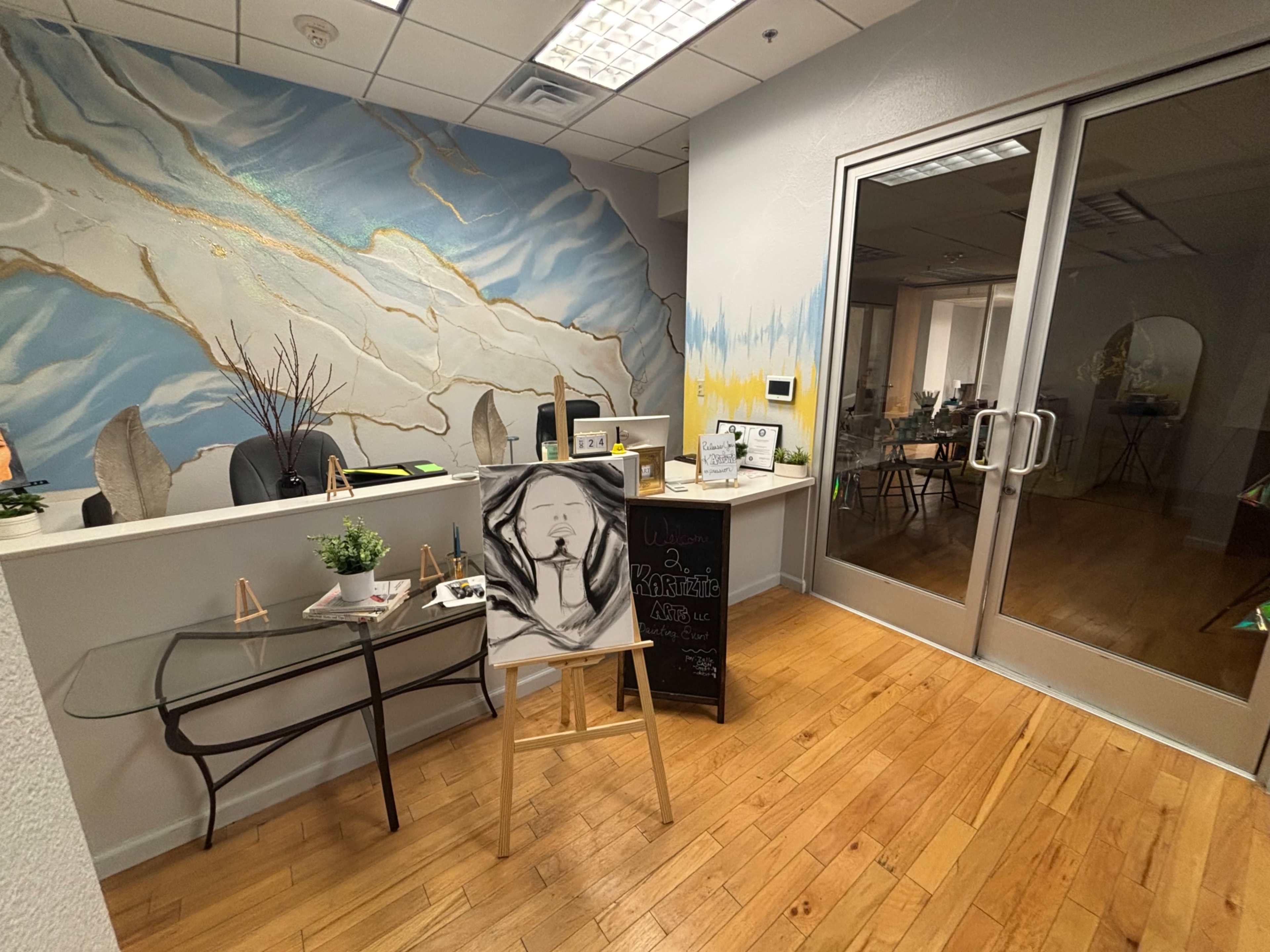 The image shows a modern reception area with a decorative mural on the wall, featuring a small table with an art piece and a glass door leading to another room.