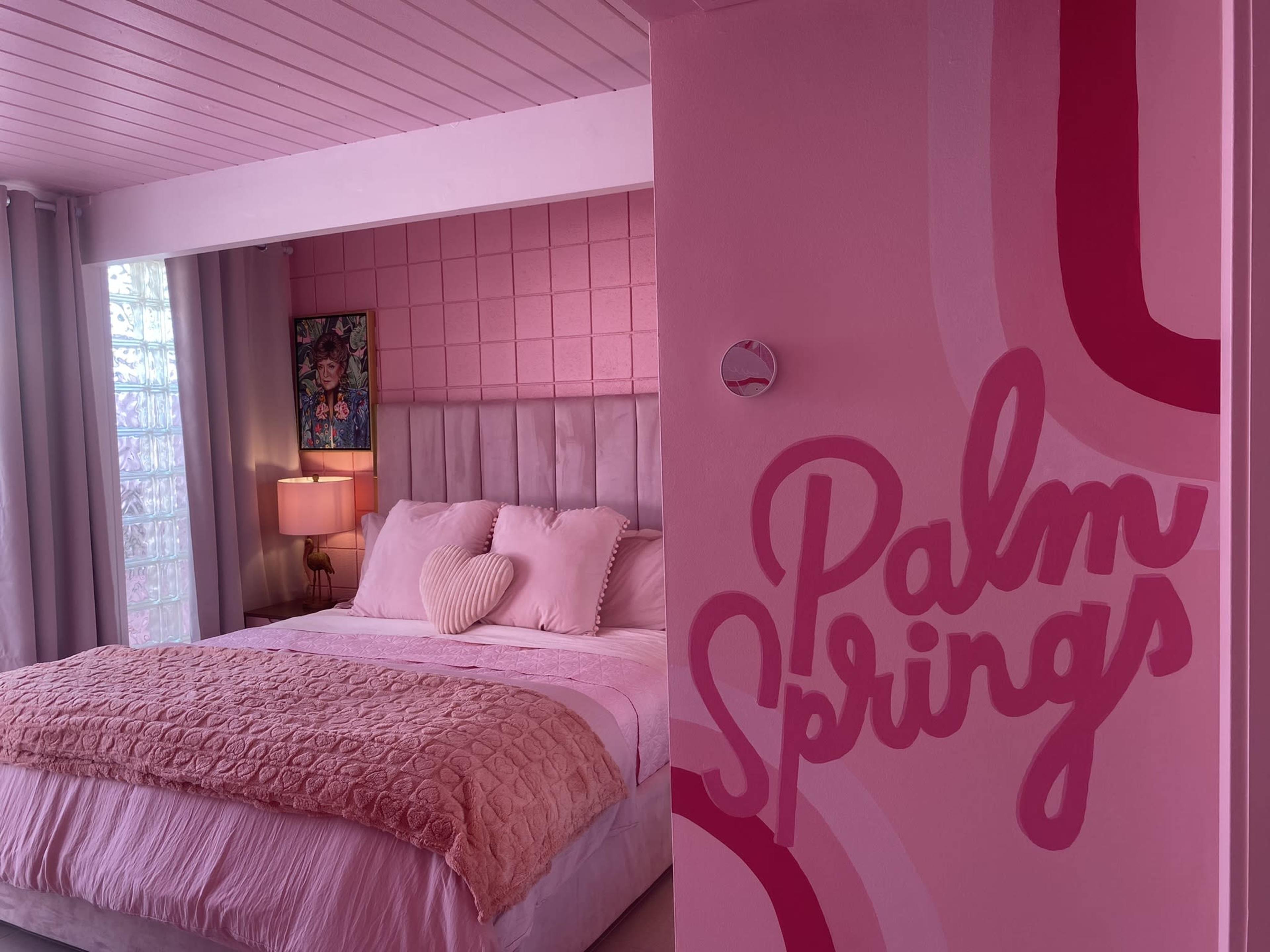 A brightly colored bedroom featuring a pink wall with "Palm Springs" written on it, a large bed with white and pink bedding, and a bedside lamp.