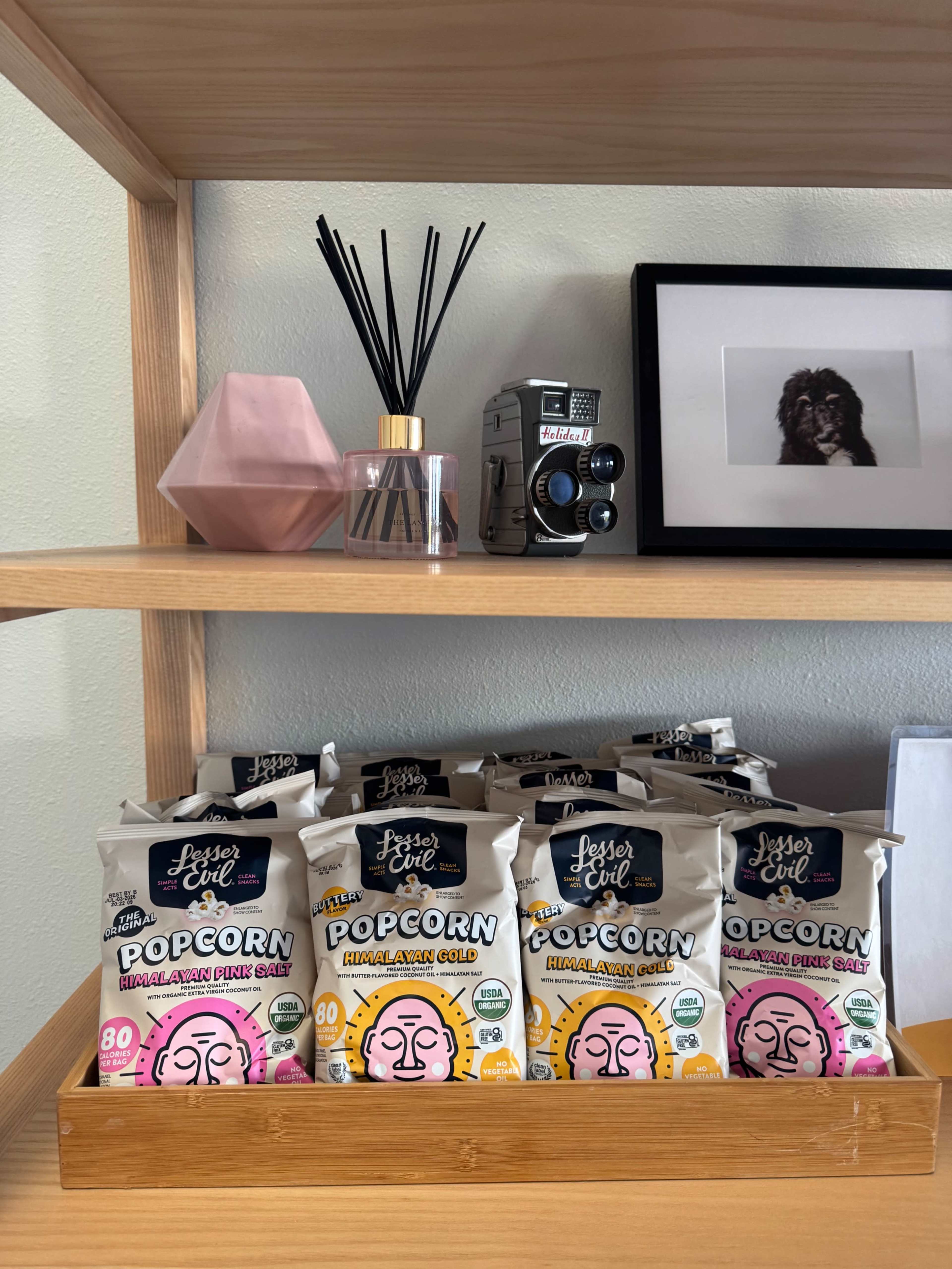 The image shows a wooden shelf displaying packages of popcorn, a pink geometric decor piece, a bottle of fragrance, an old camera, and a framed photo.