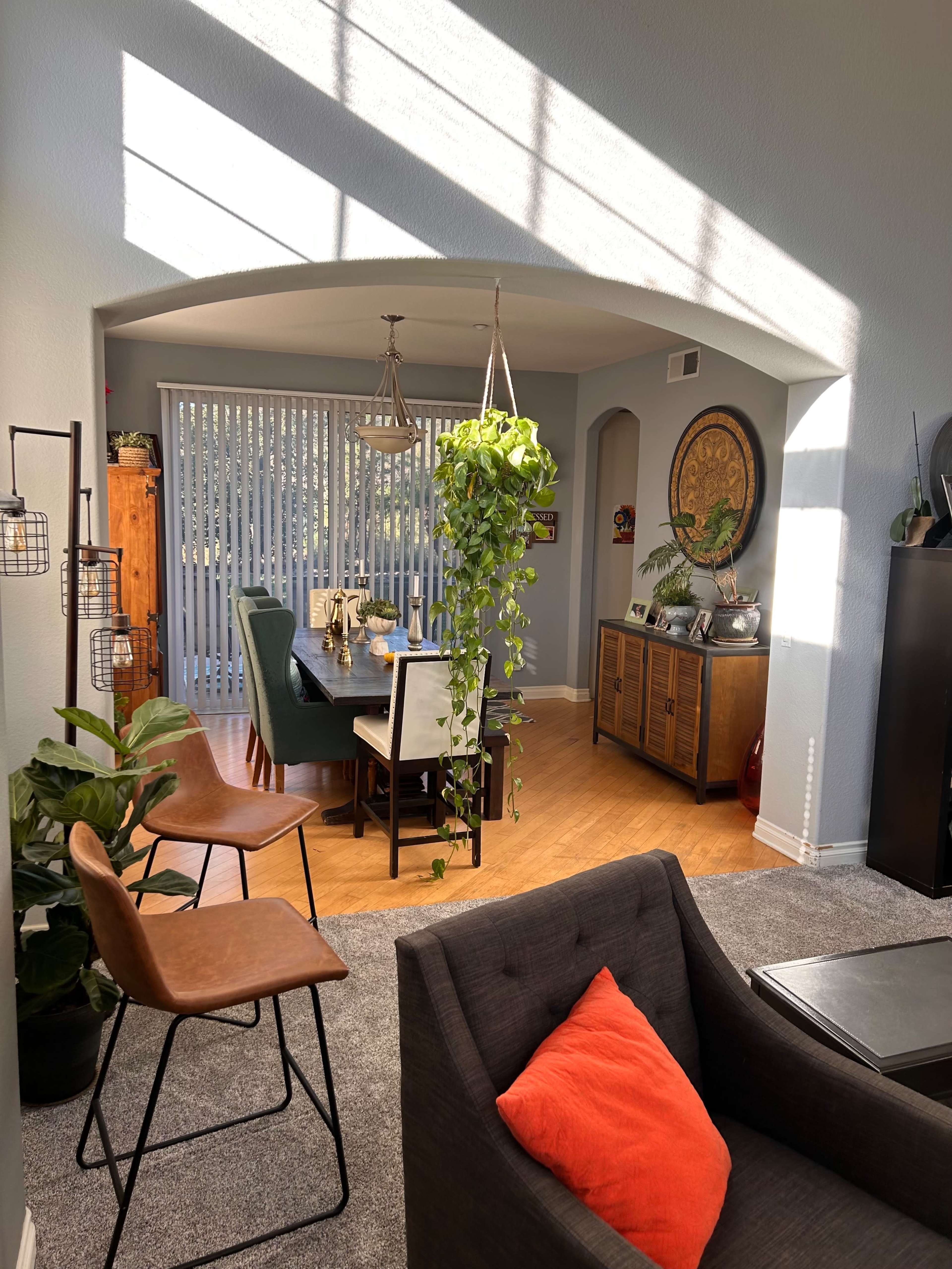 A well-lit dining area features a table surrounded by chairs, with a hanging plant and a cozy seating space visible in the foreground.