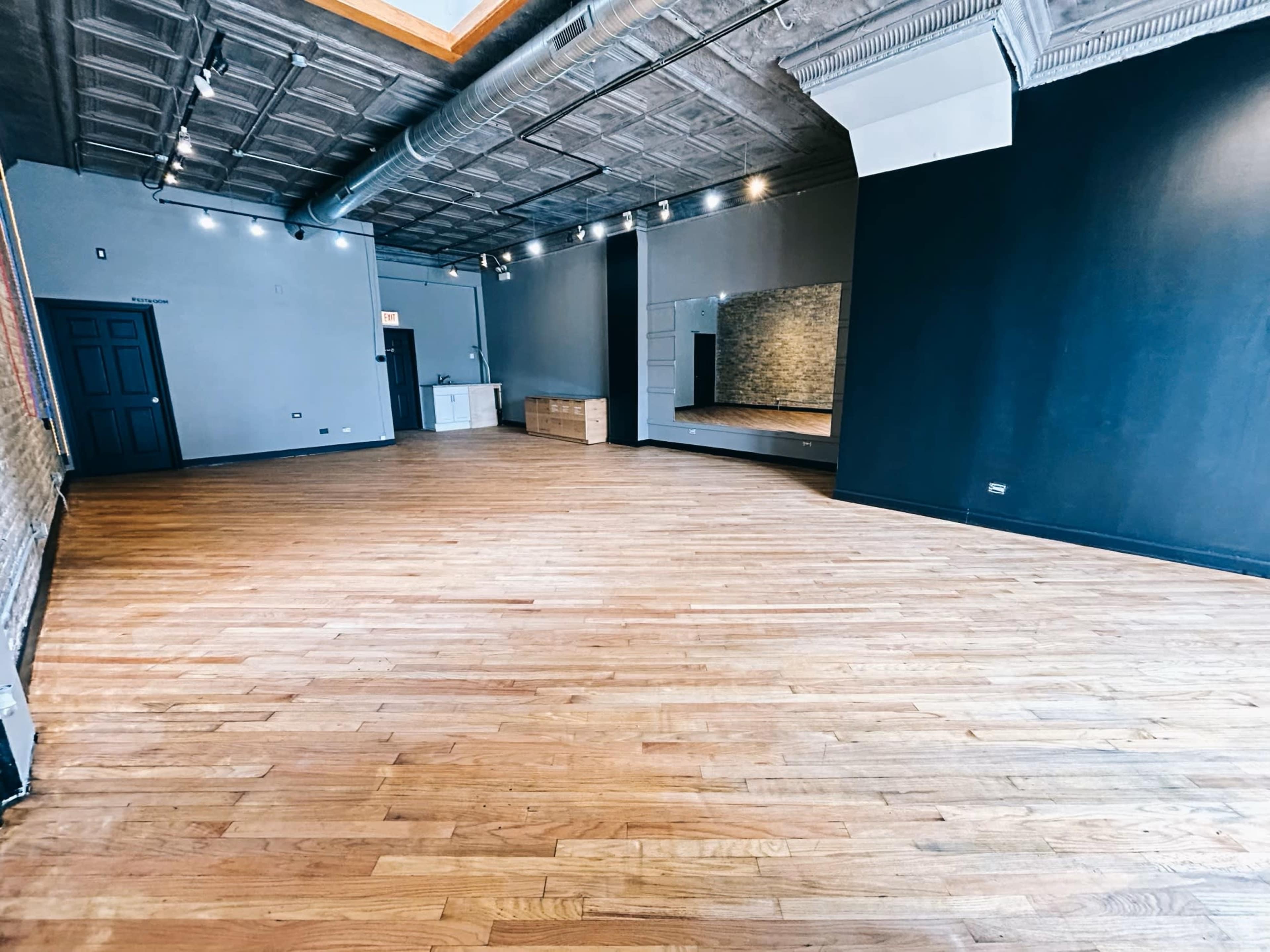 The image shows an empty studio space with polished wooden floors, exposed brick walls, and a large mirror on one side.