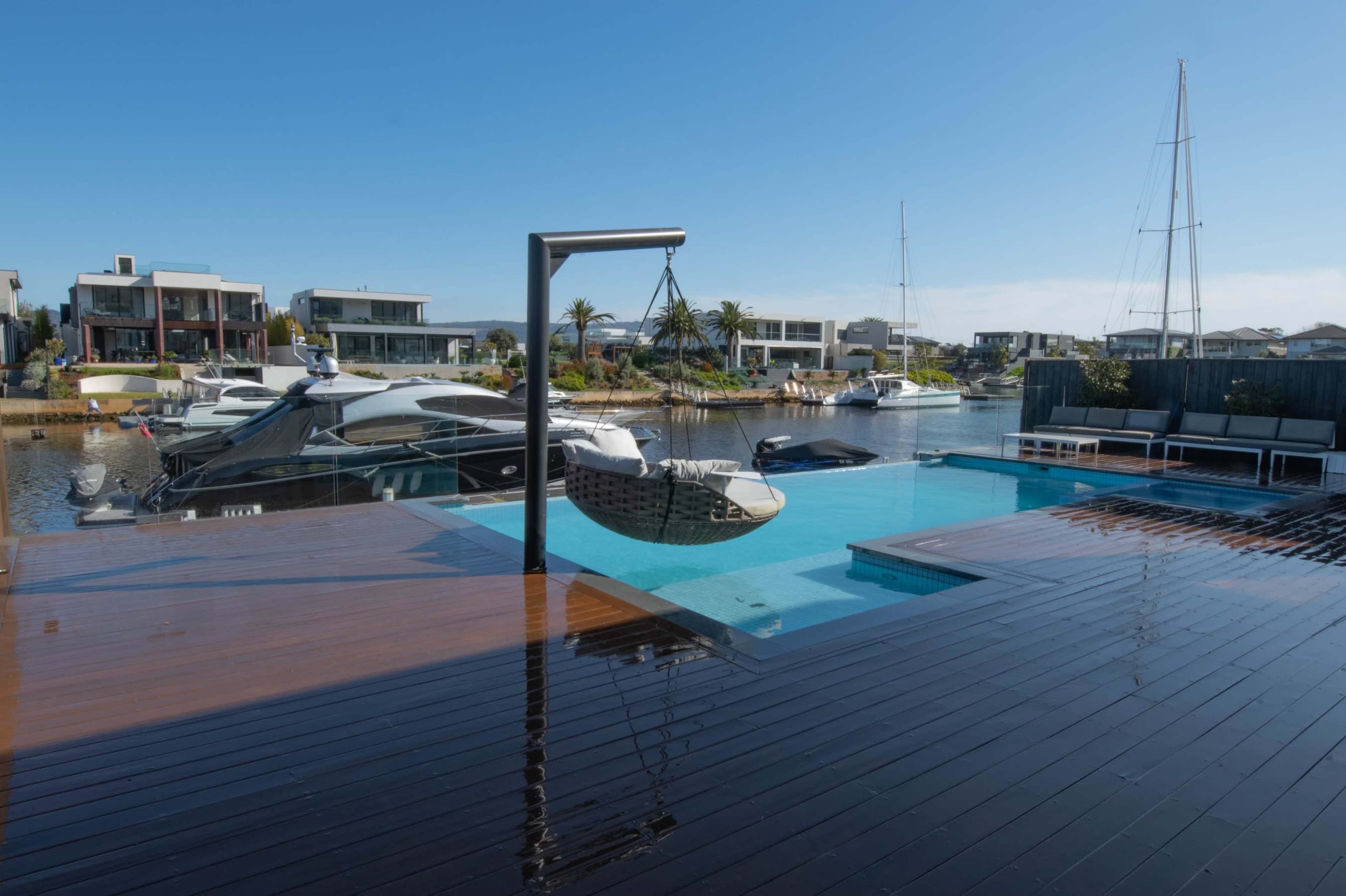 A modern waterfront property with a swimming pool, a hammock on a stand, and boats docked in a canal lined with houses.
