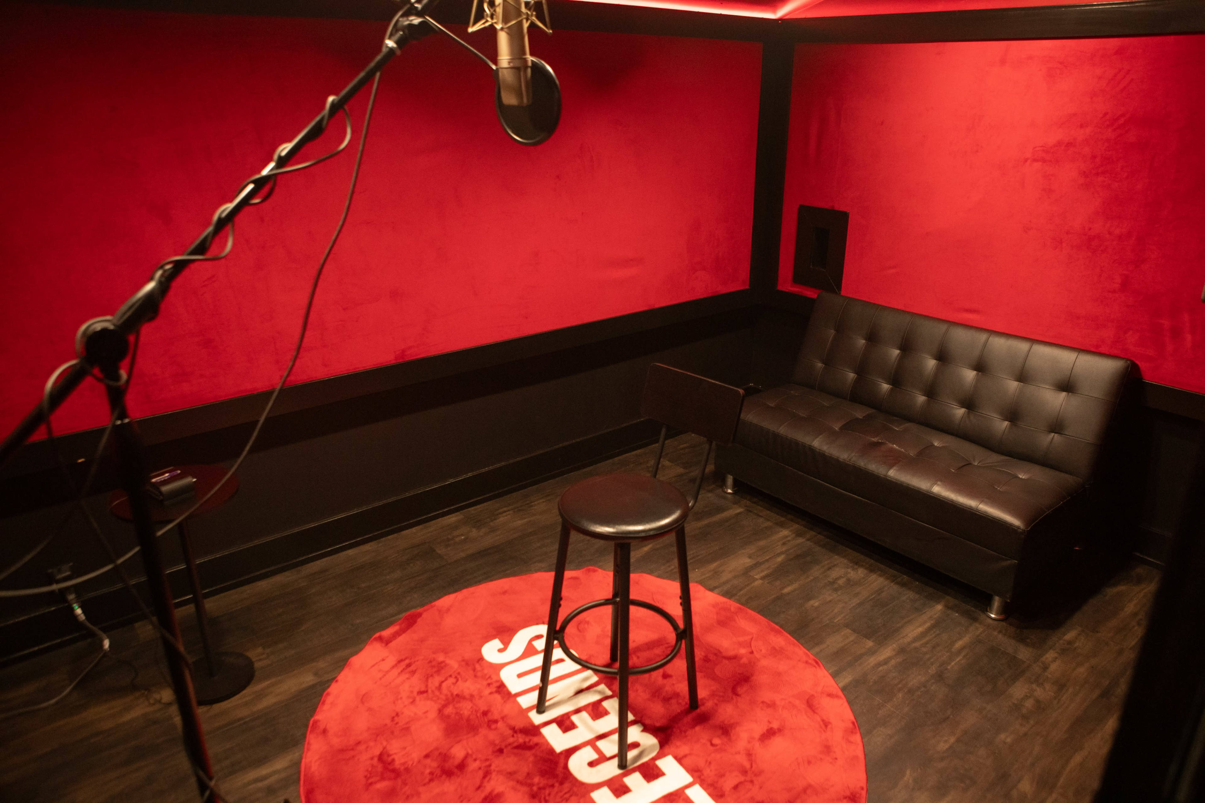 The image shows a small recording studio with red walls, a microphone on a stand, a black sofa, and a stool on a round red rug.