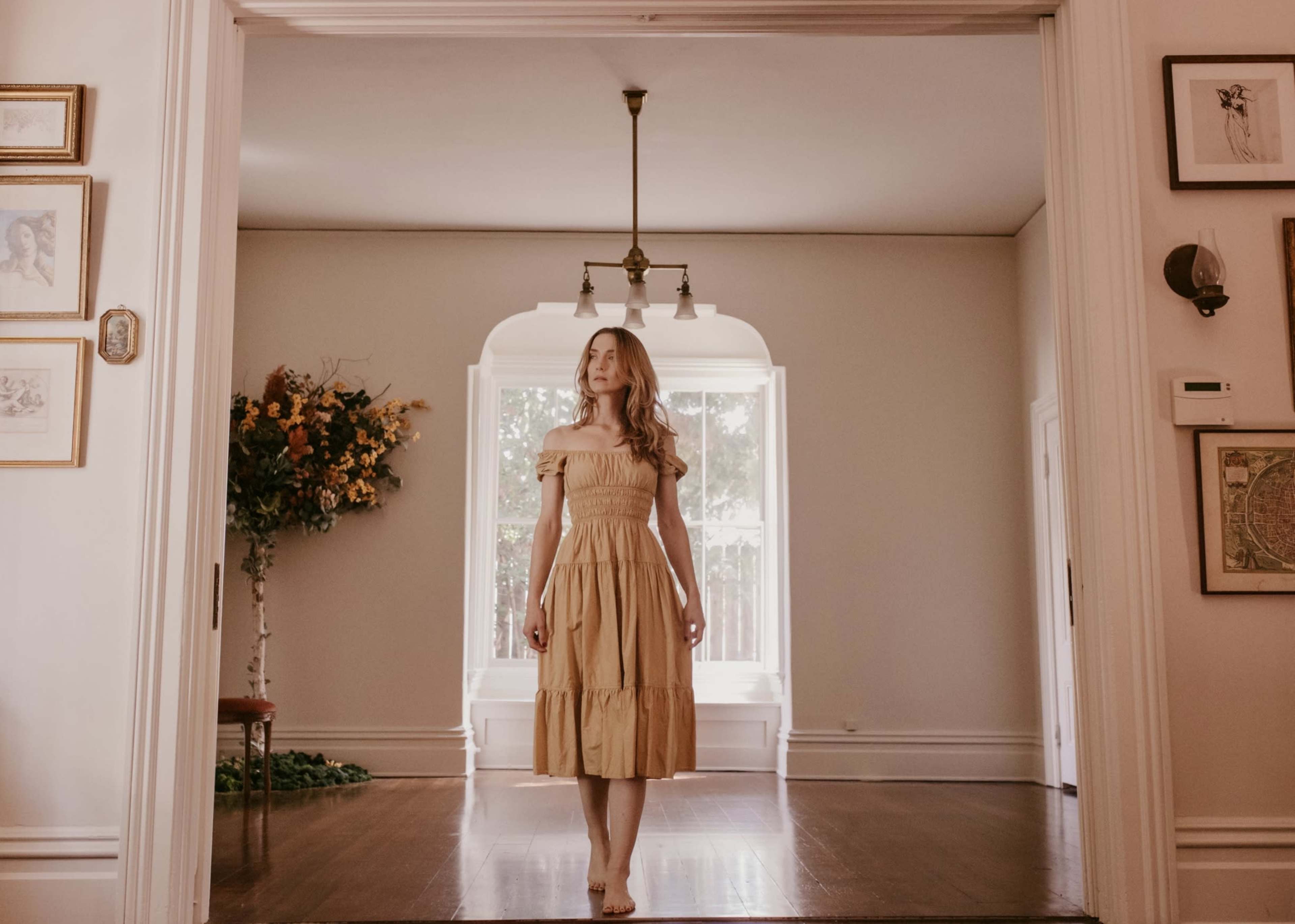 A woman in a yellow off-the-shoulder dress stands in the doorway of a light-filled room adorned with framed artwork and a large window.
